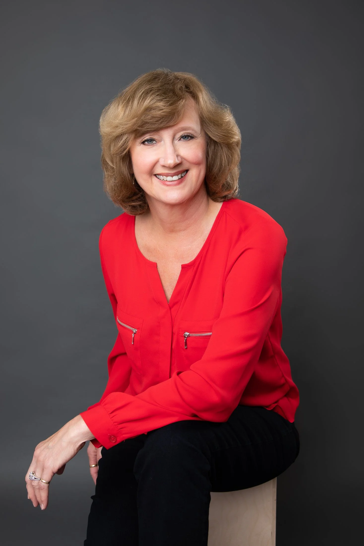 Headshot of a woman with wavy blonde hair and green eyes, wearing a red blouse on a medium gray background. Photographed in studio by Amanda Faucett Photography.