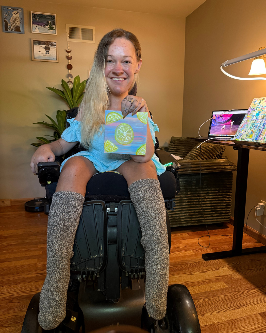 A woman in a power wheelchair holding a colorful painting of lemon slices, smiling in a cozy living room with wooden floors, potted plant, artwork on the wall, and a laptop on a nearby stand.