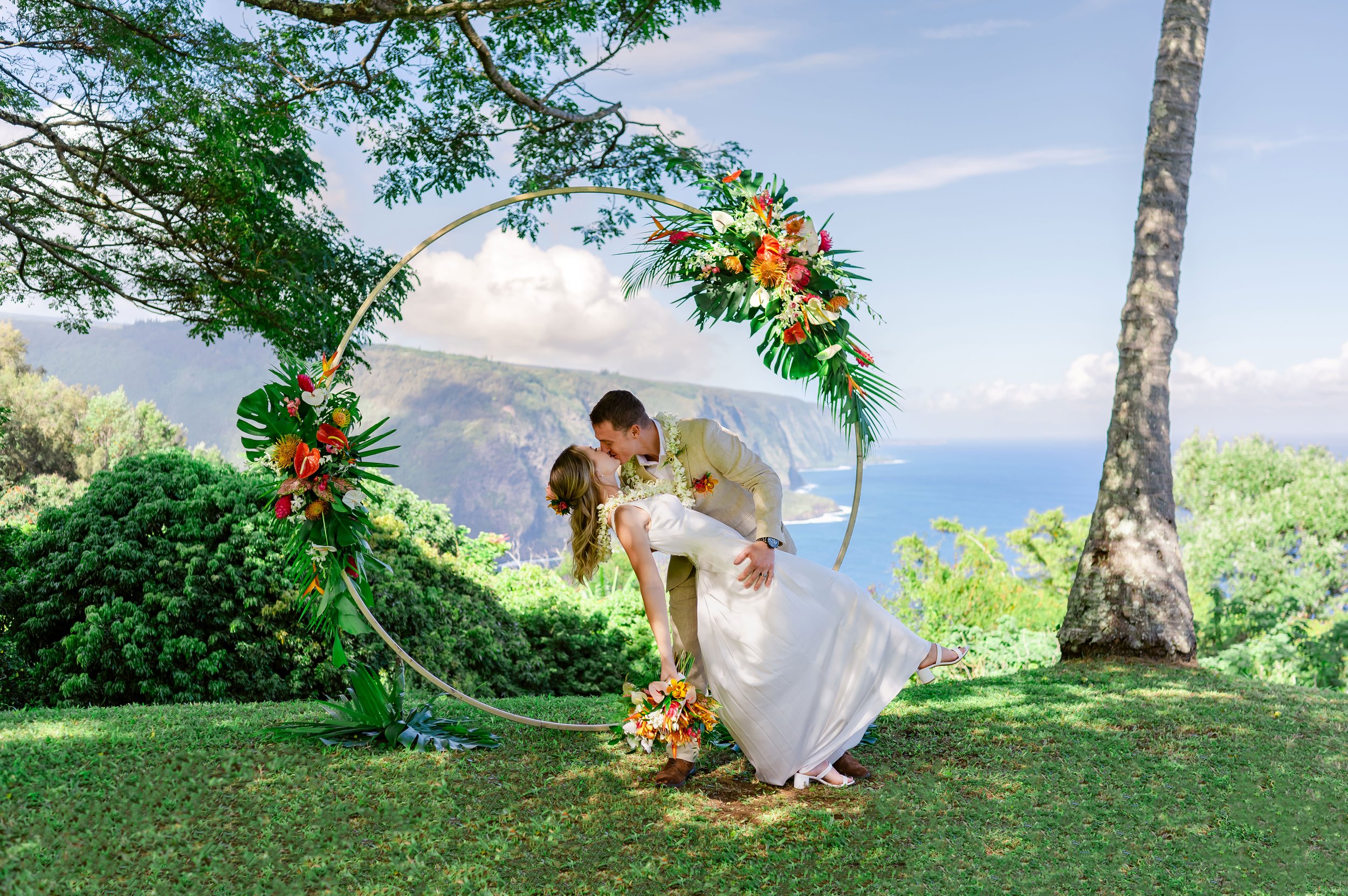 Bride and groom kissing under a floral arch, with tropical scenery in the background, including ocean cliffs and lush greenery.
