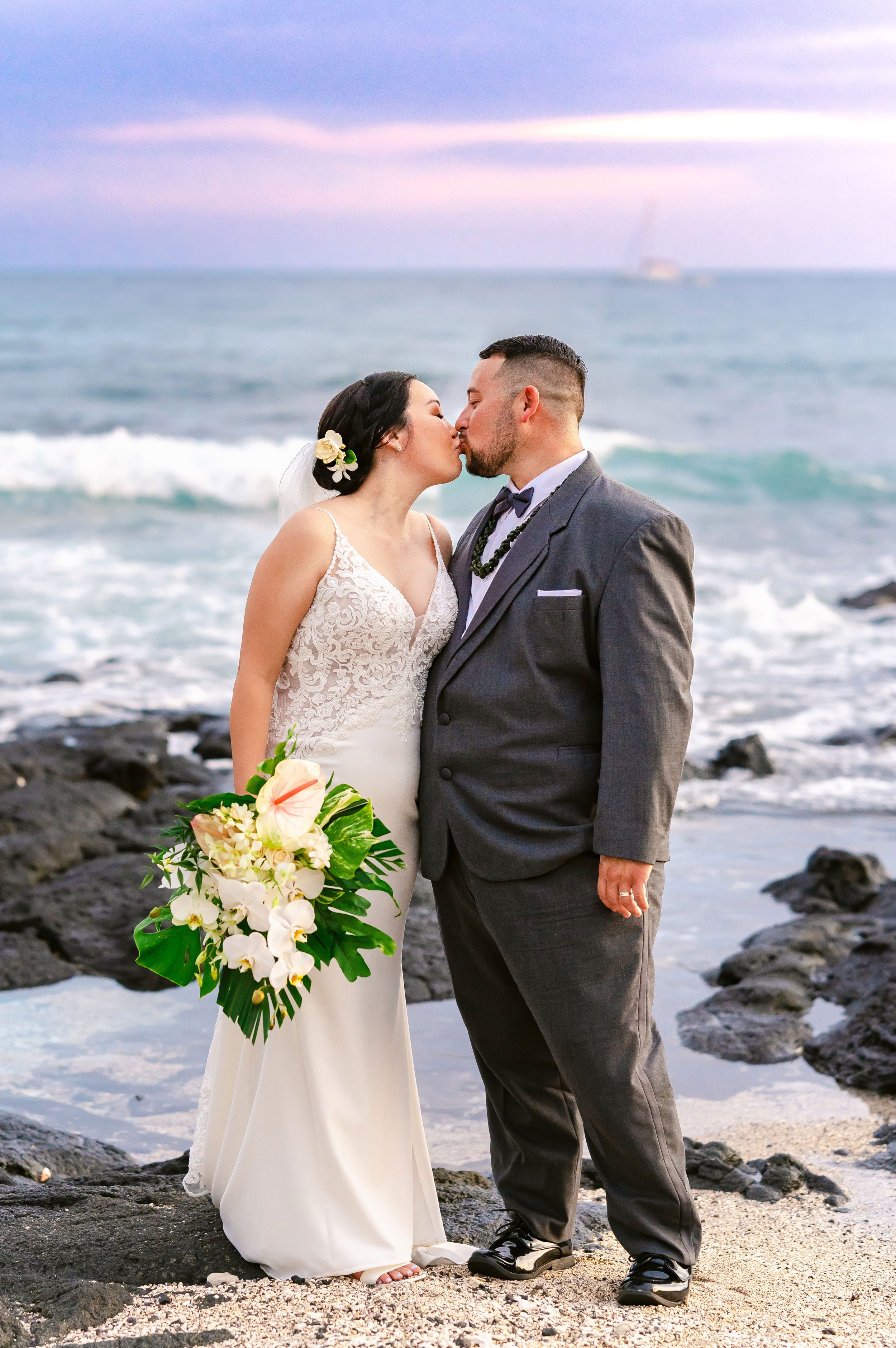 A couple kissing on a rocky beach during a wedding, with the ocean and a colorful sky in the background. The bride is wearing a white lace dress and holding a bouquet, while the groom is in a gray suit.