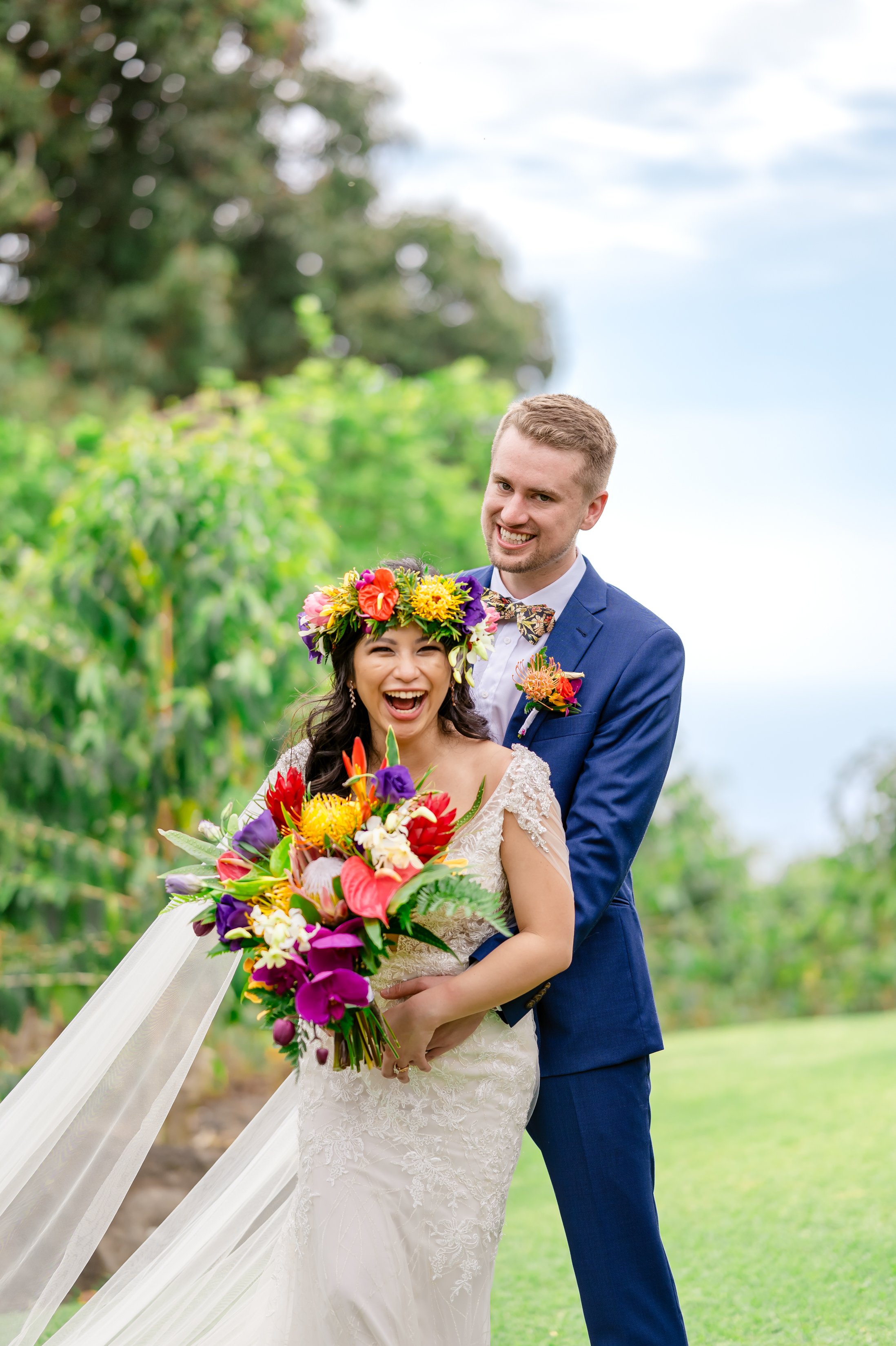 A bride and groom smiling at their outdoor wedding, with the bride wearing a floral crown and holding a colorful bouquet, and the groom wearing a blue suit.