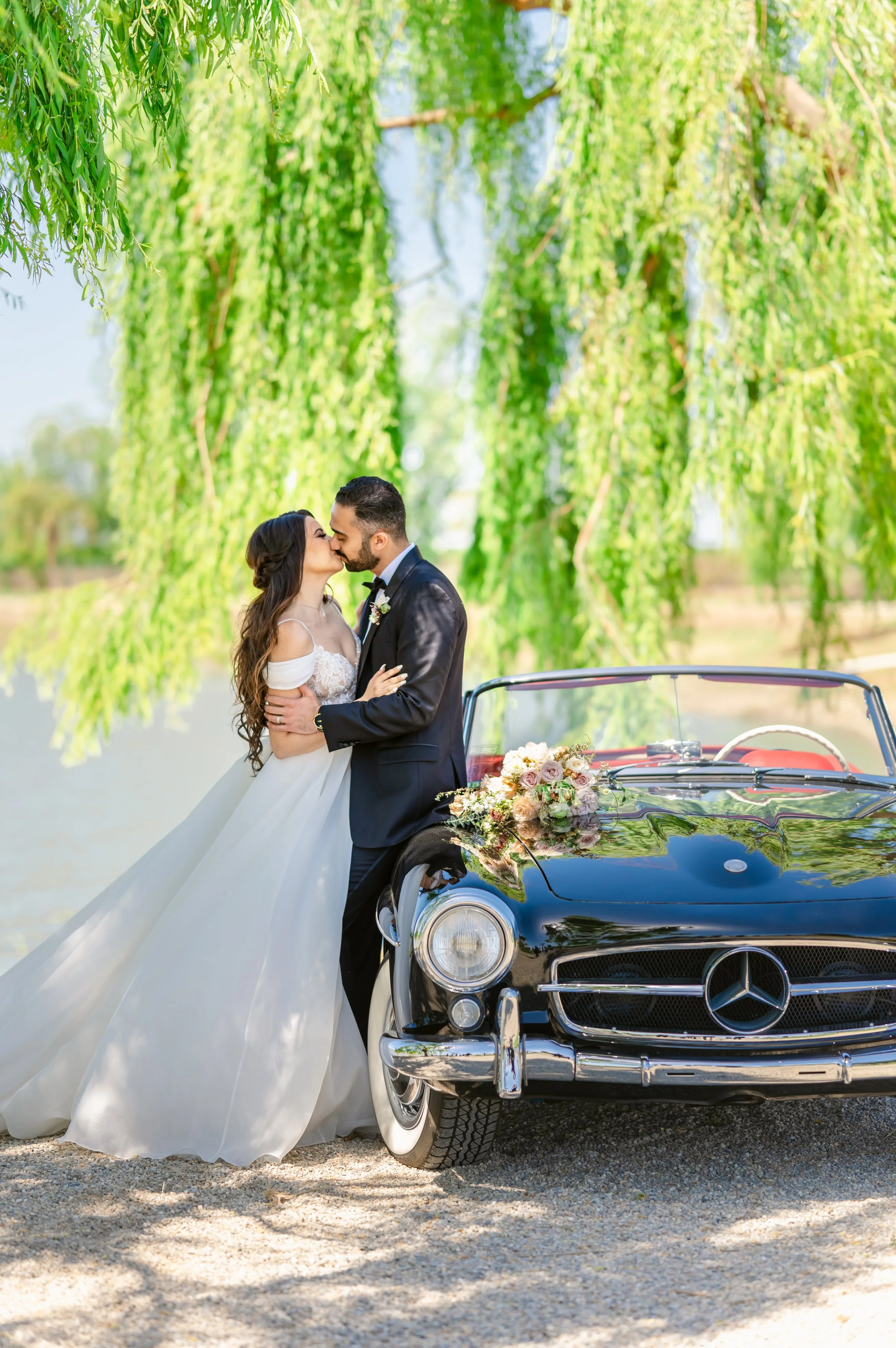 Bride and groom kissing in front of a vintage black car with floral decorations, under a lush green tree, near a body of water.