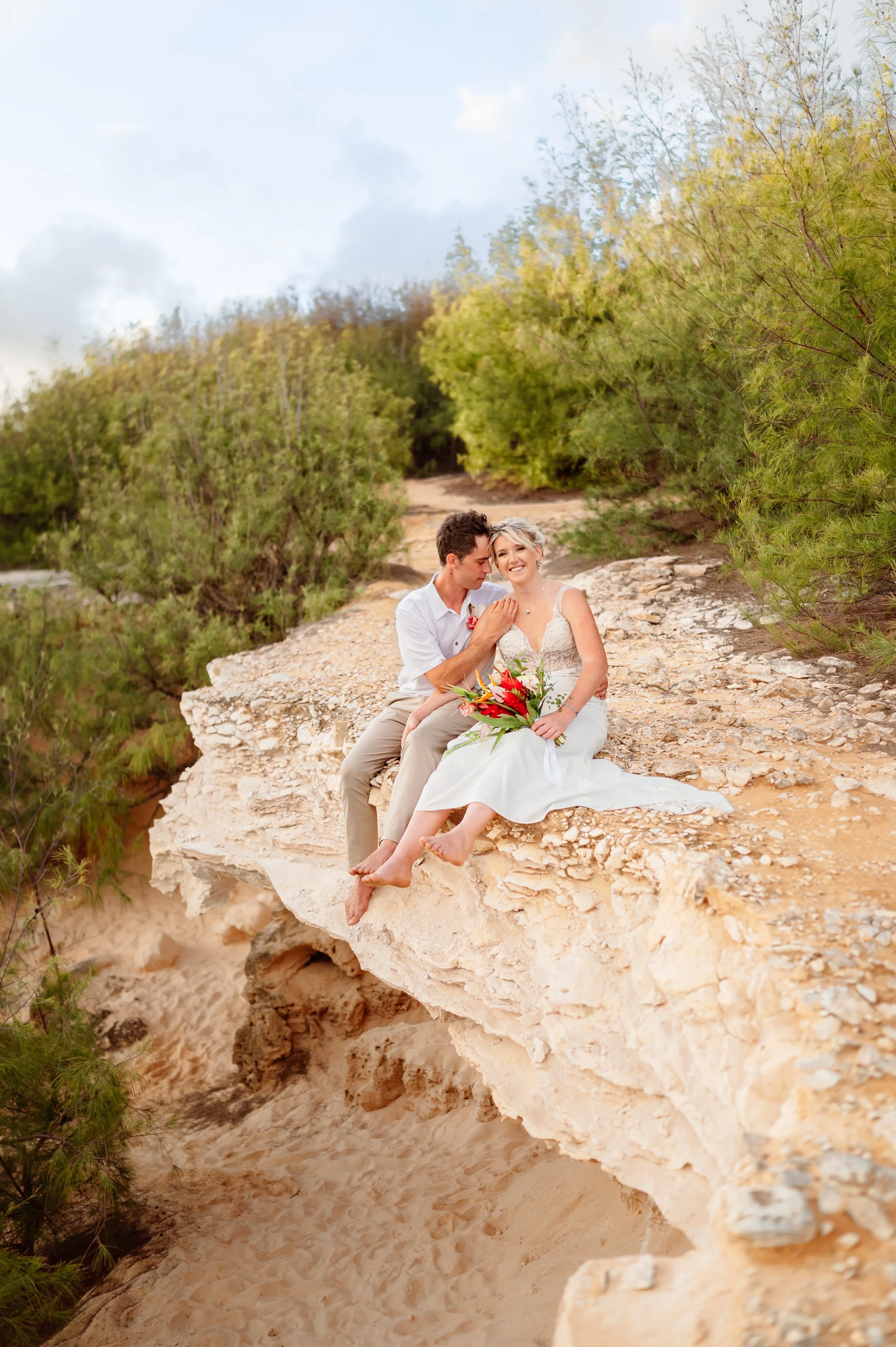 Couple sitting on a rocky ledge, woman in wedding dress holding a bouquet, man in casual attire, surrounded by greenery.
