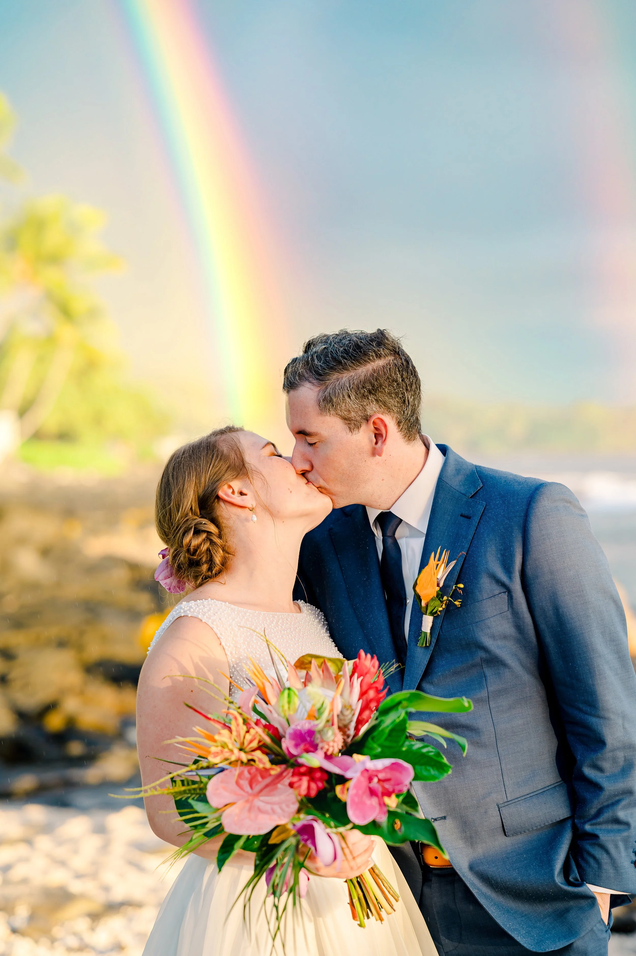 Couple kissing on a beach with a rainbow in the background, holding a bouquet of colorful flowers.