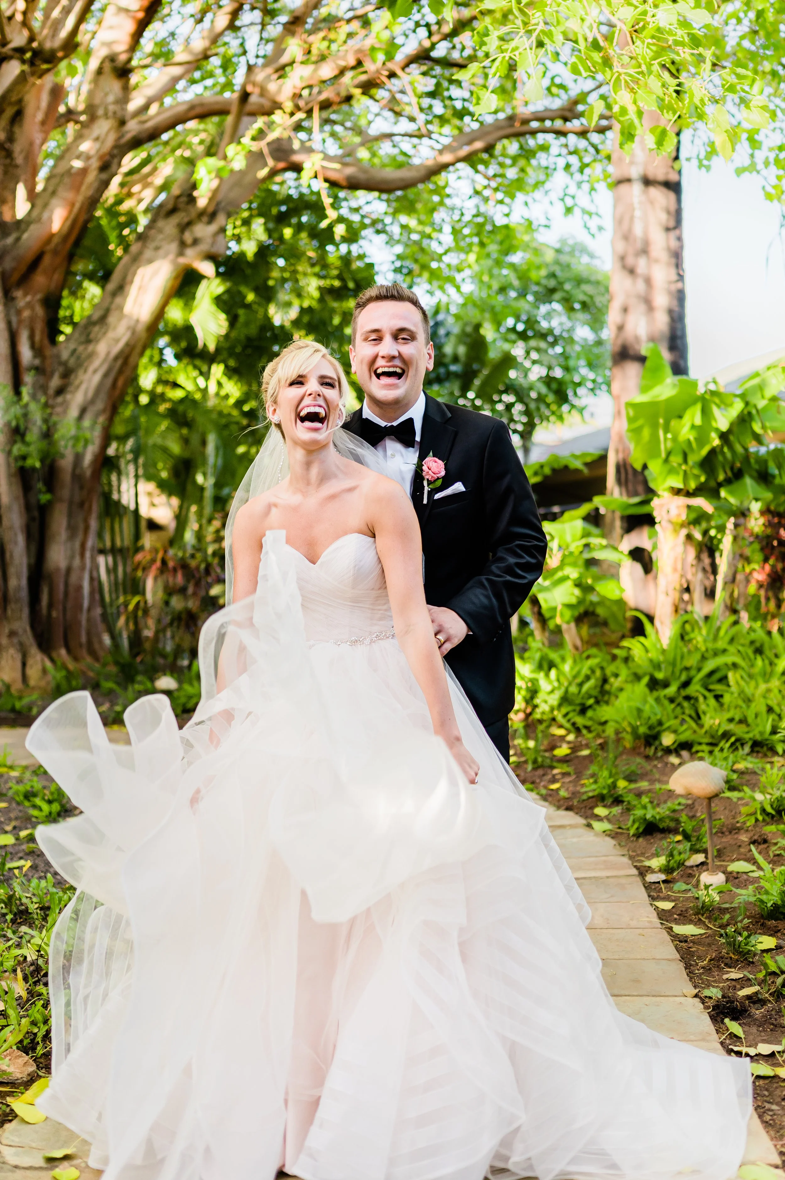 Bride and groom laughing outdoors, under a tree, in wedding attire.