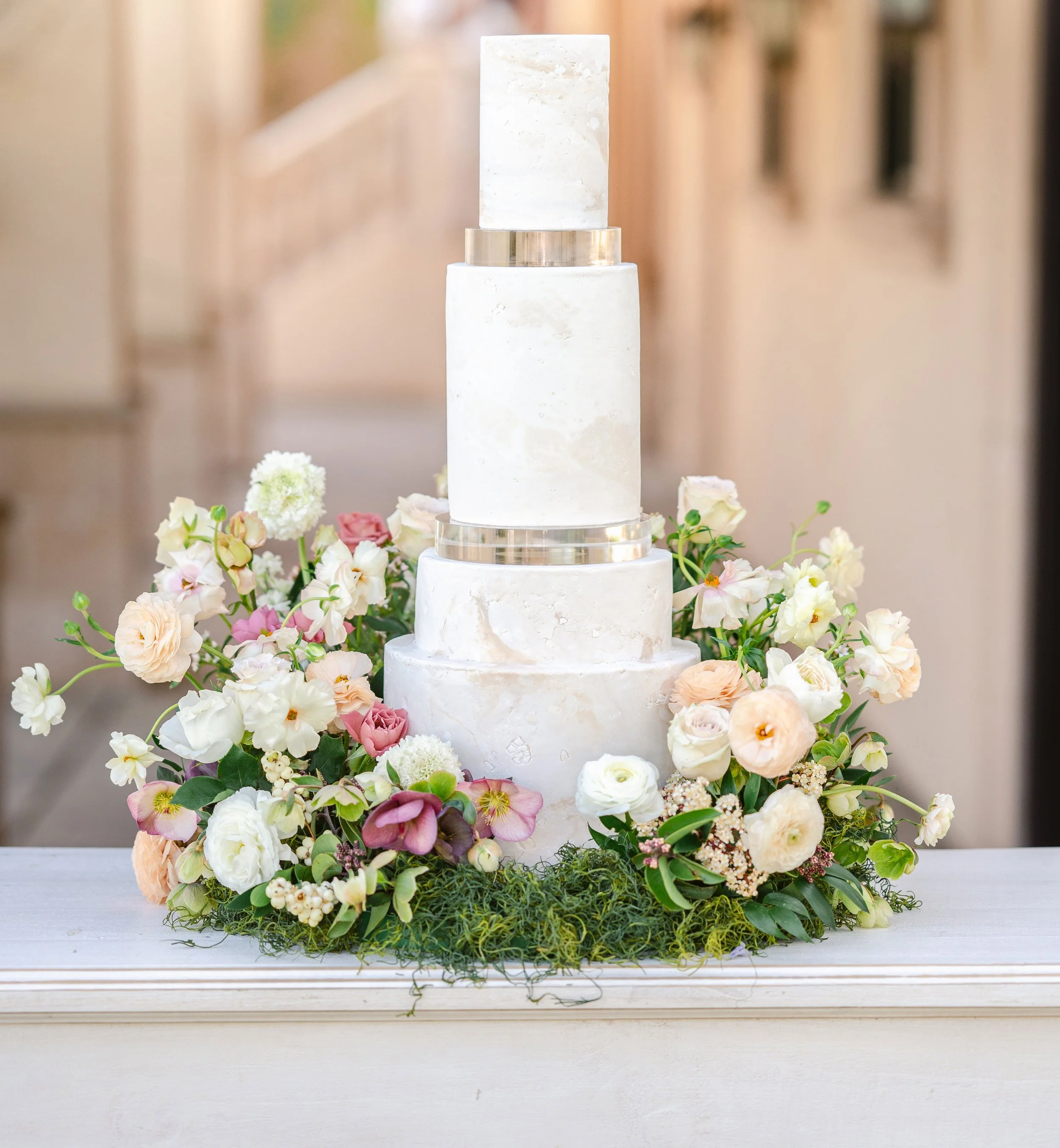 Four-tier white wedding cake with metallic accents, surrounded by an arrangement of pastel flowers and greenery on a table.