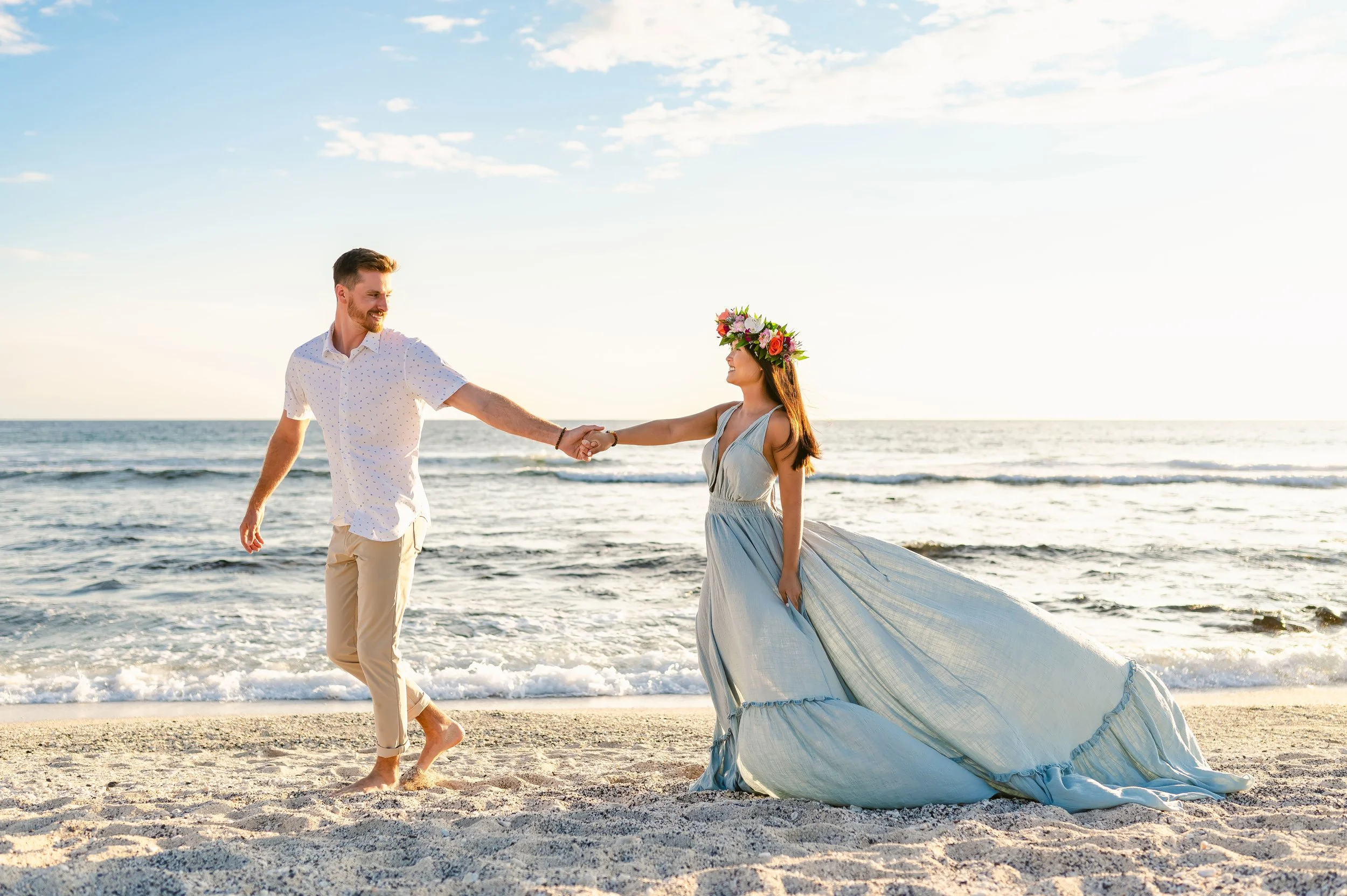 Couple walking hand in hand on beach, woman in light blue dress with floral crown, sunny day with ocean background.