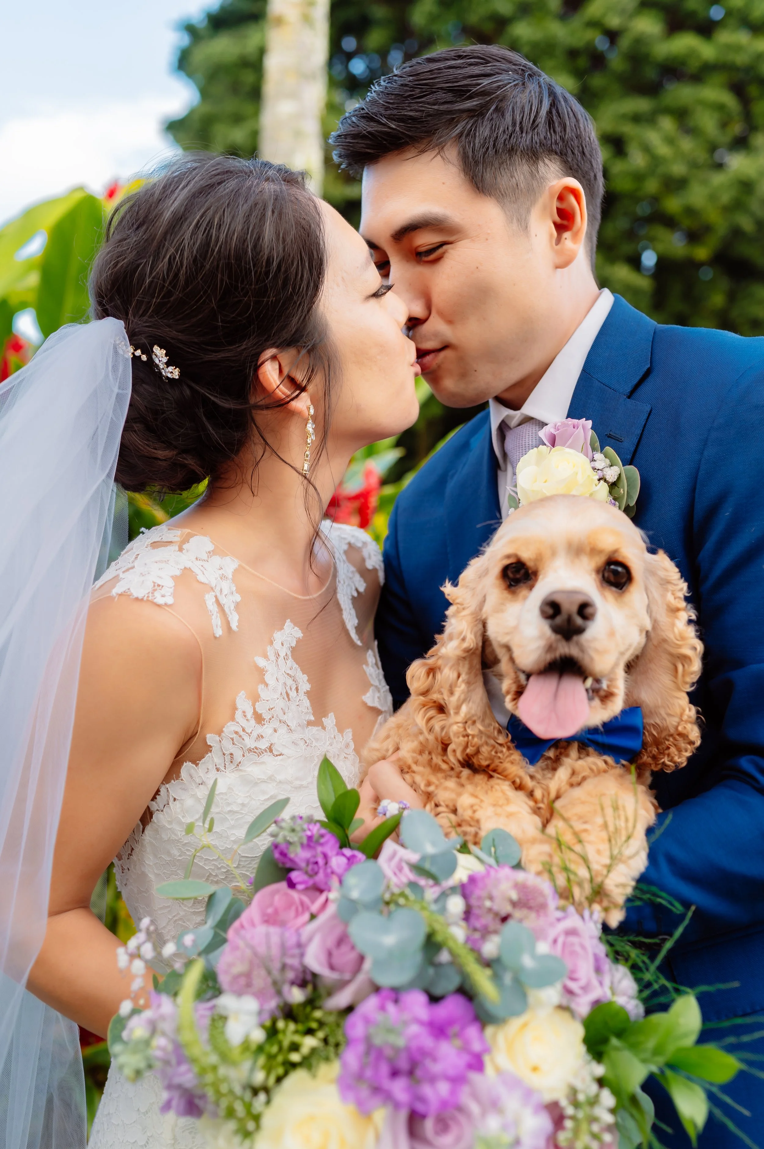 Bride and groom kissing, holding a dog, with a bouquet of flowers.