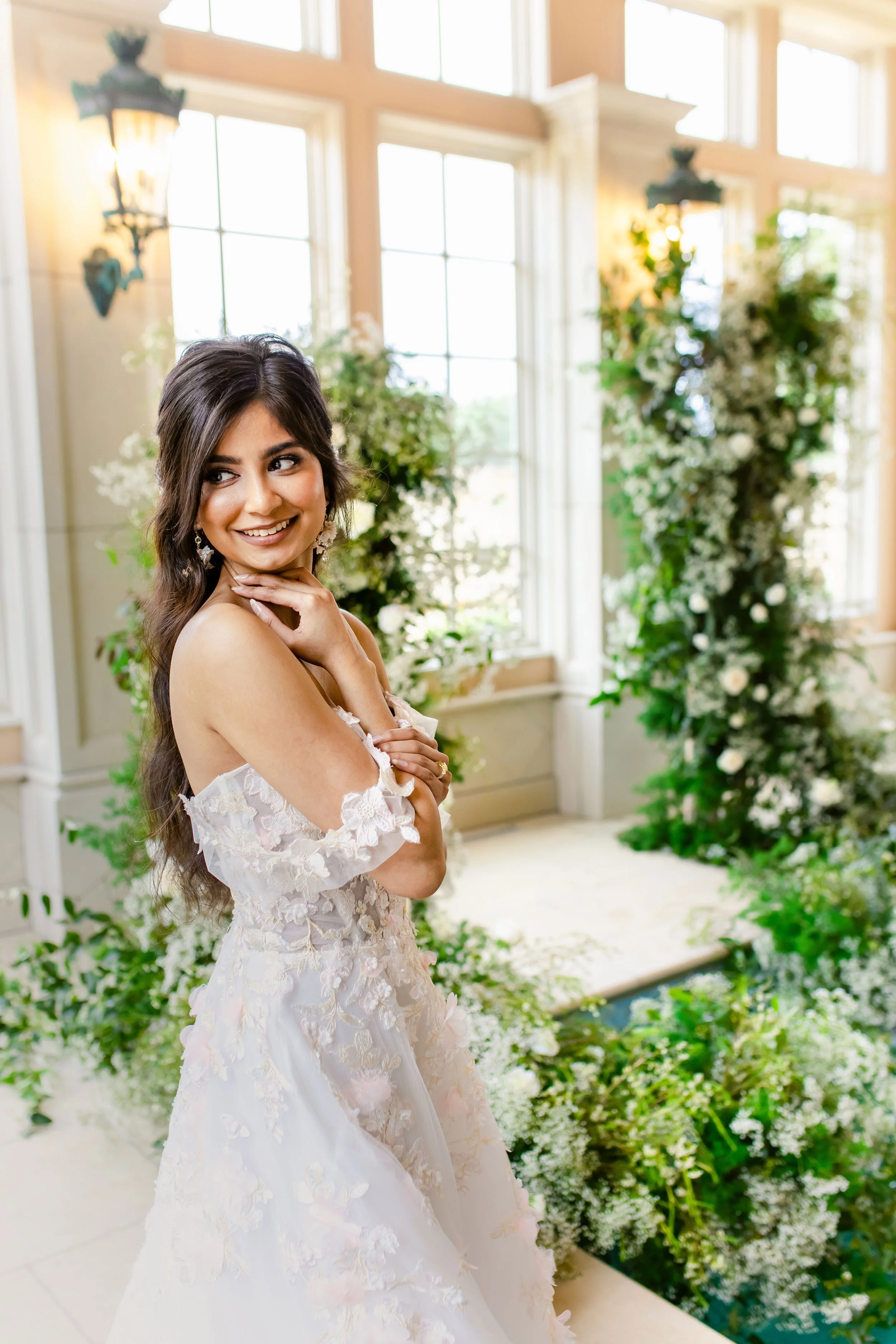 A woman in a floral lace dress standing inside a sunlit room with large windows, surrounded by greenery and floral decorations.