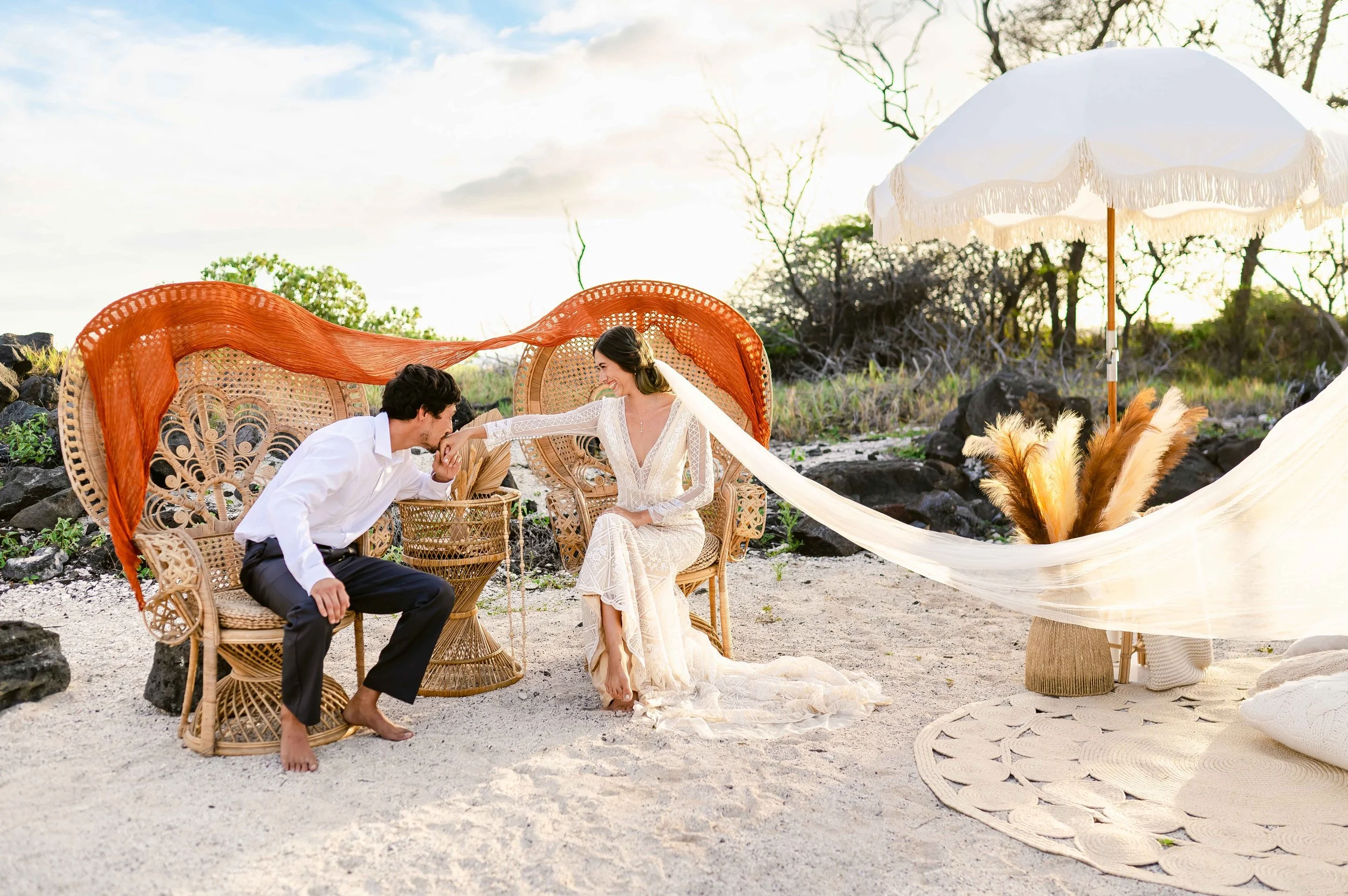 Couple sitting on wicker chairs at a beach wedding, with man kissing woman's hand. Decor includes parasol, pampas grass, and neutral tones.