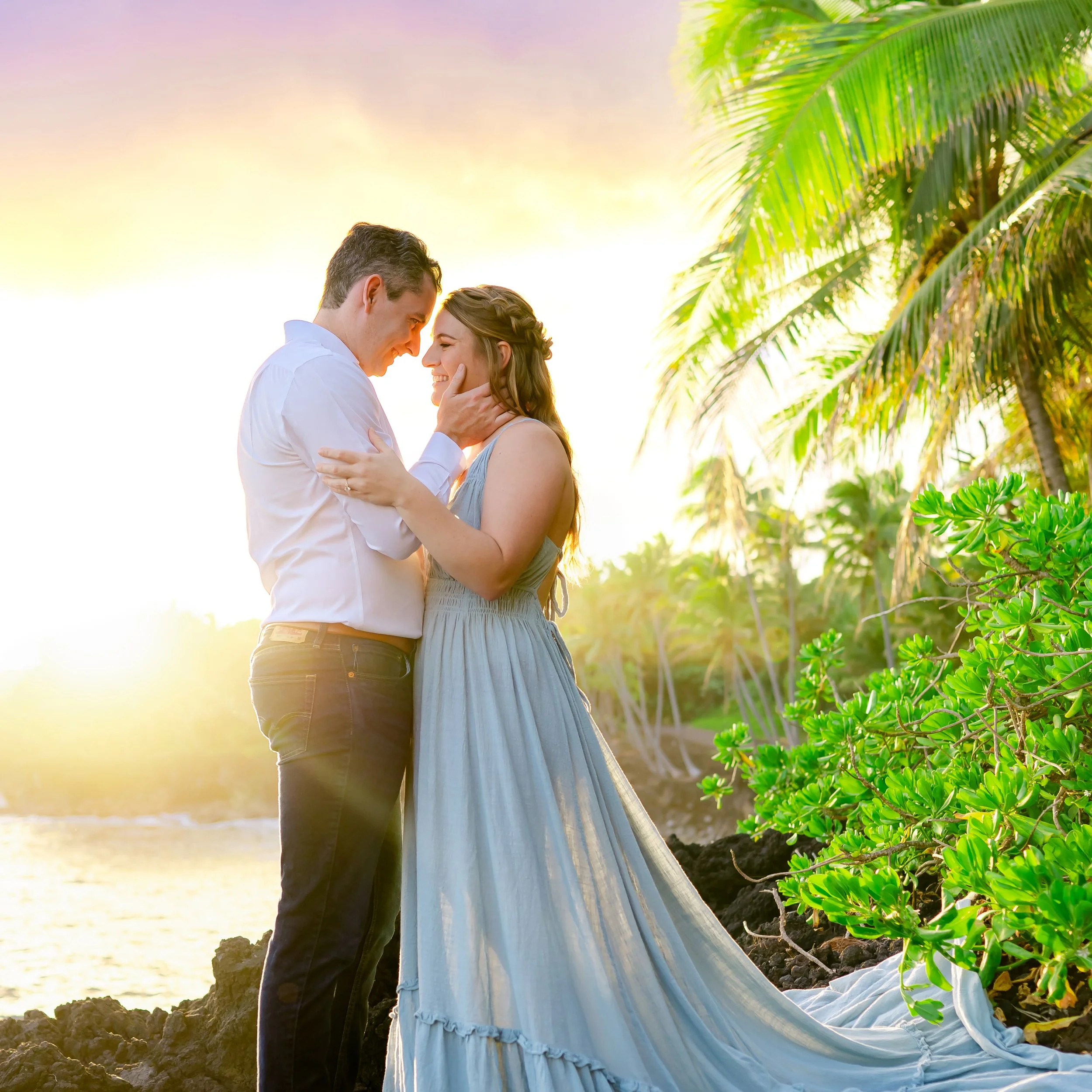 A couple embracing near the ocean with palm trees in the background at sunset.