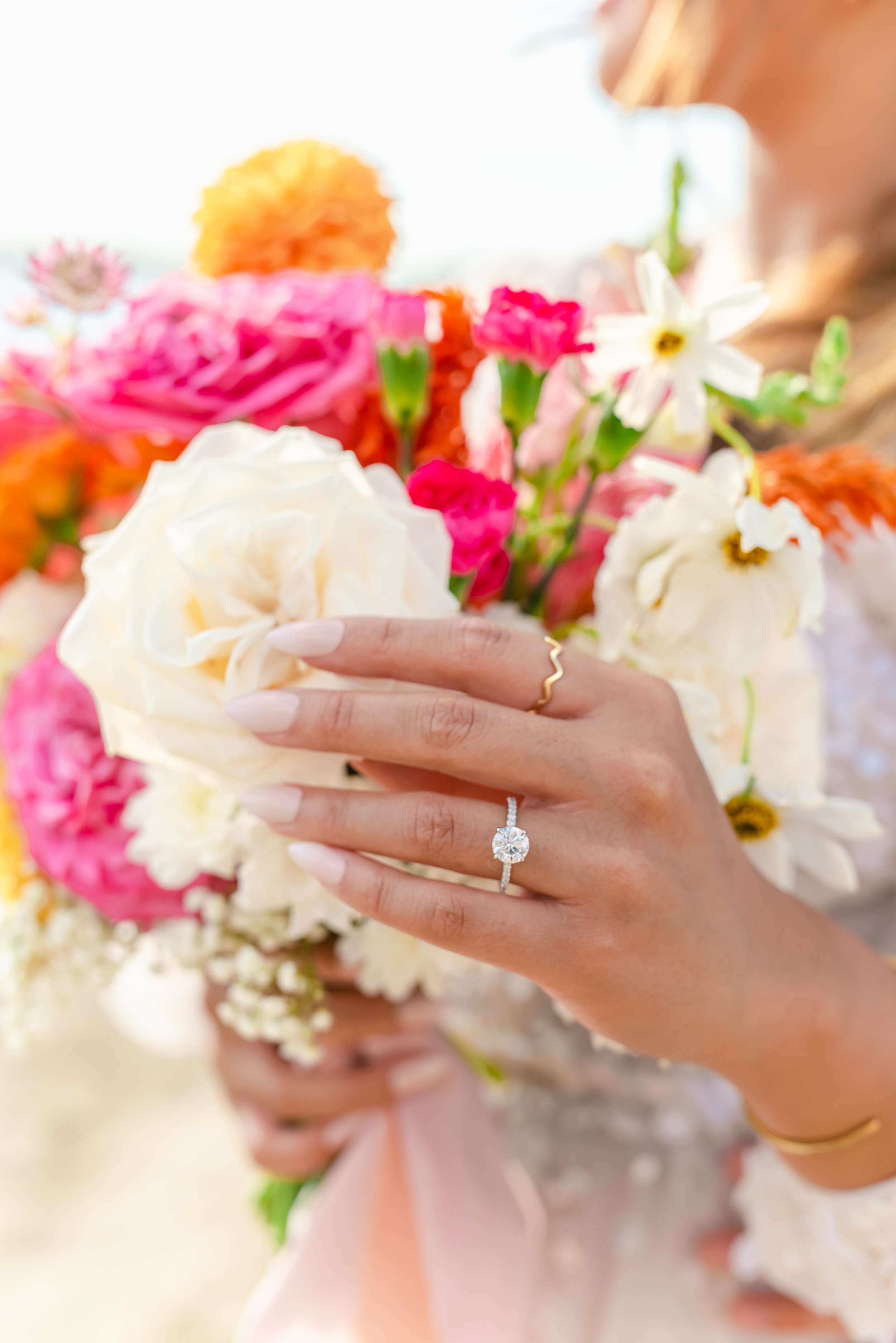 Close-up of a hand with a diamond engagement ring holding a bouquet of colorful flowers, including roses and daisies.