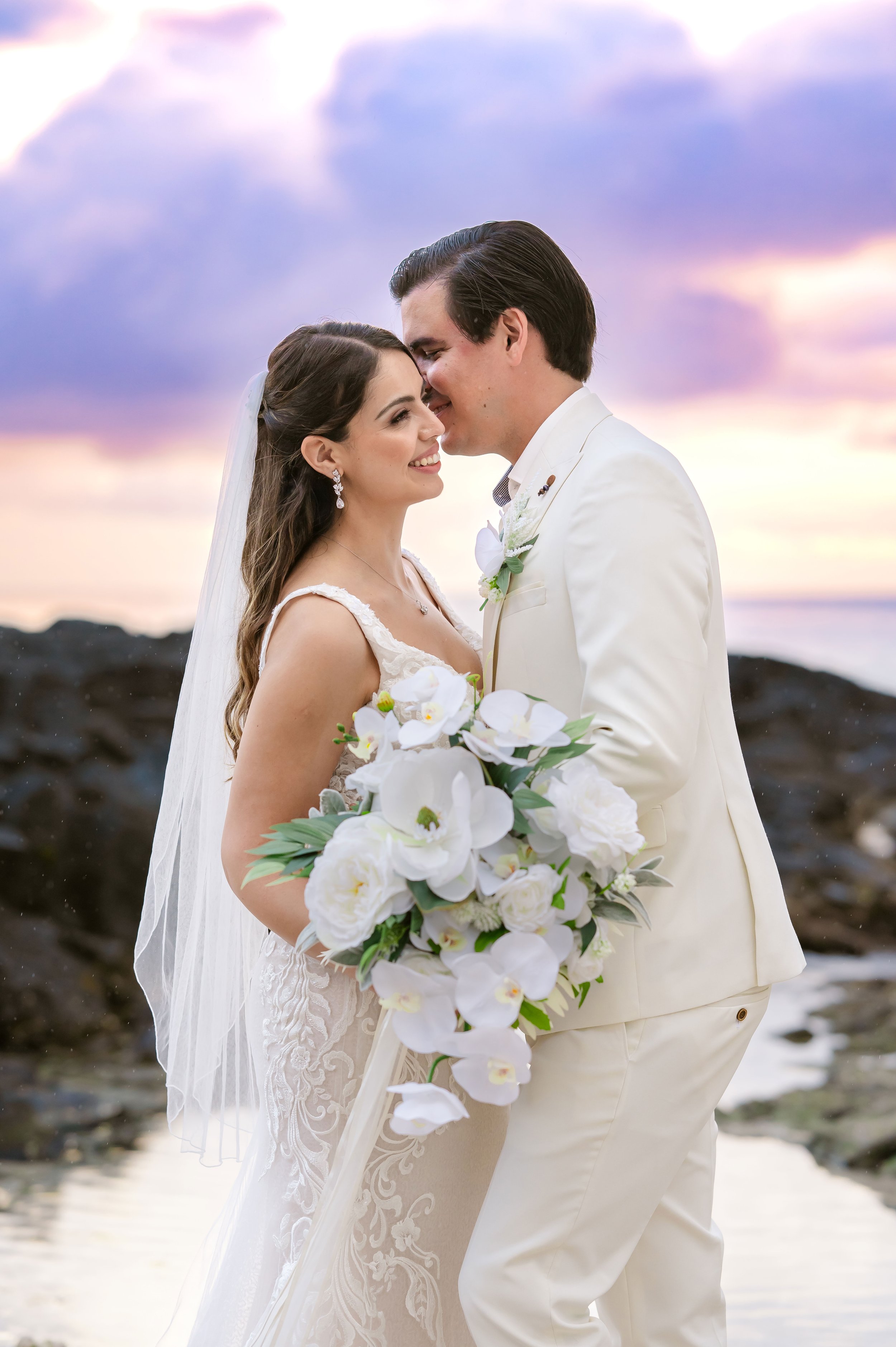 Bride and groom at beach wedding with sunset backdrop, bride holding white bouquet, both dressed in white.