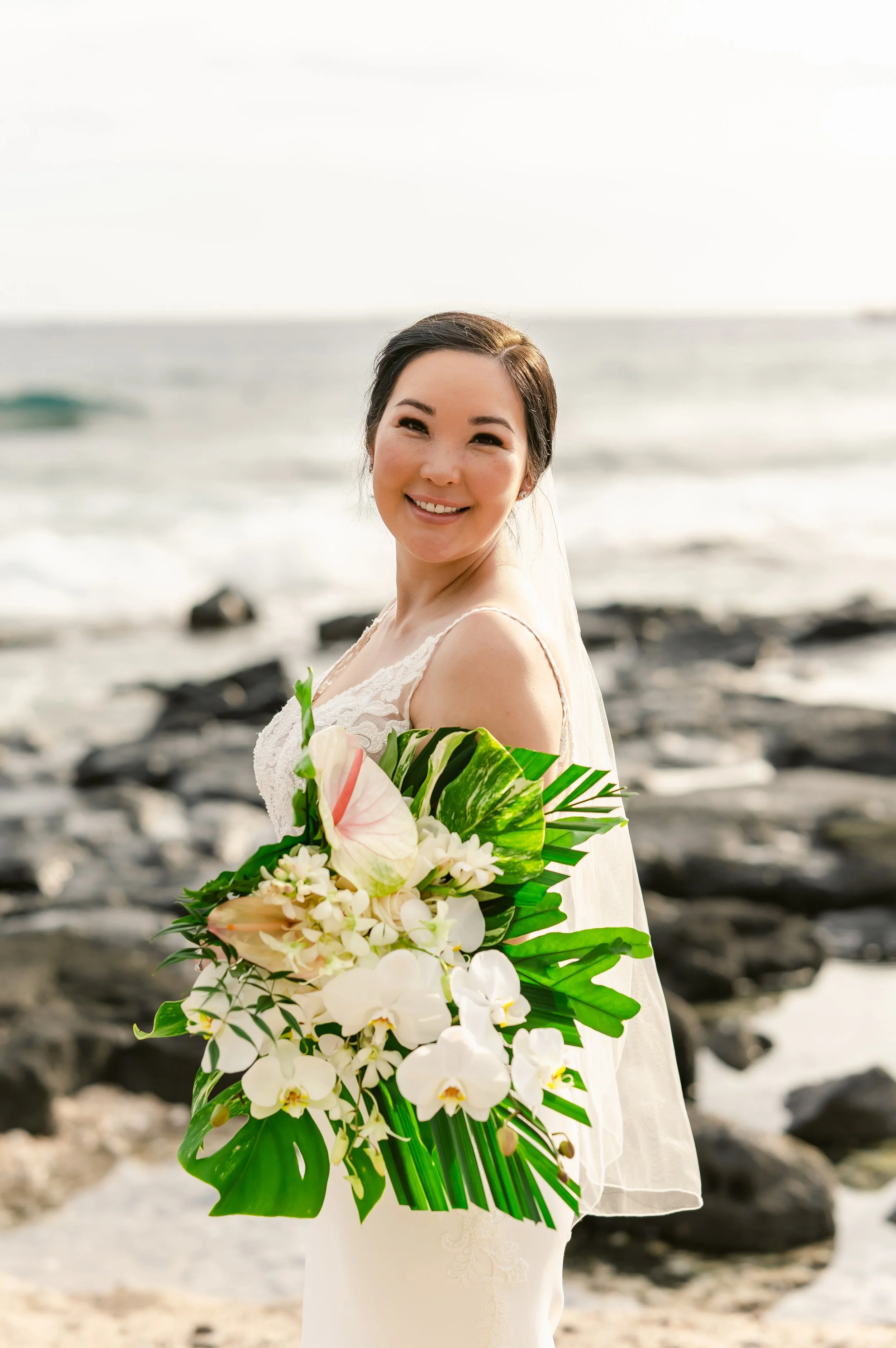 Bride on beach with tropical bouquet, wearing veil and white dress, ocean in background.