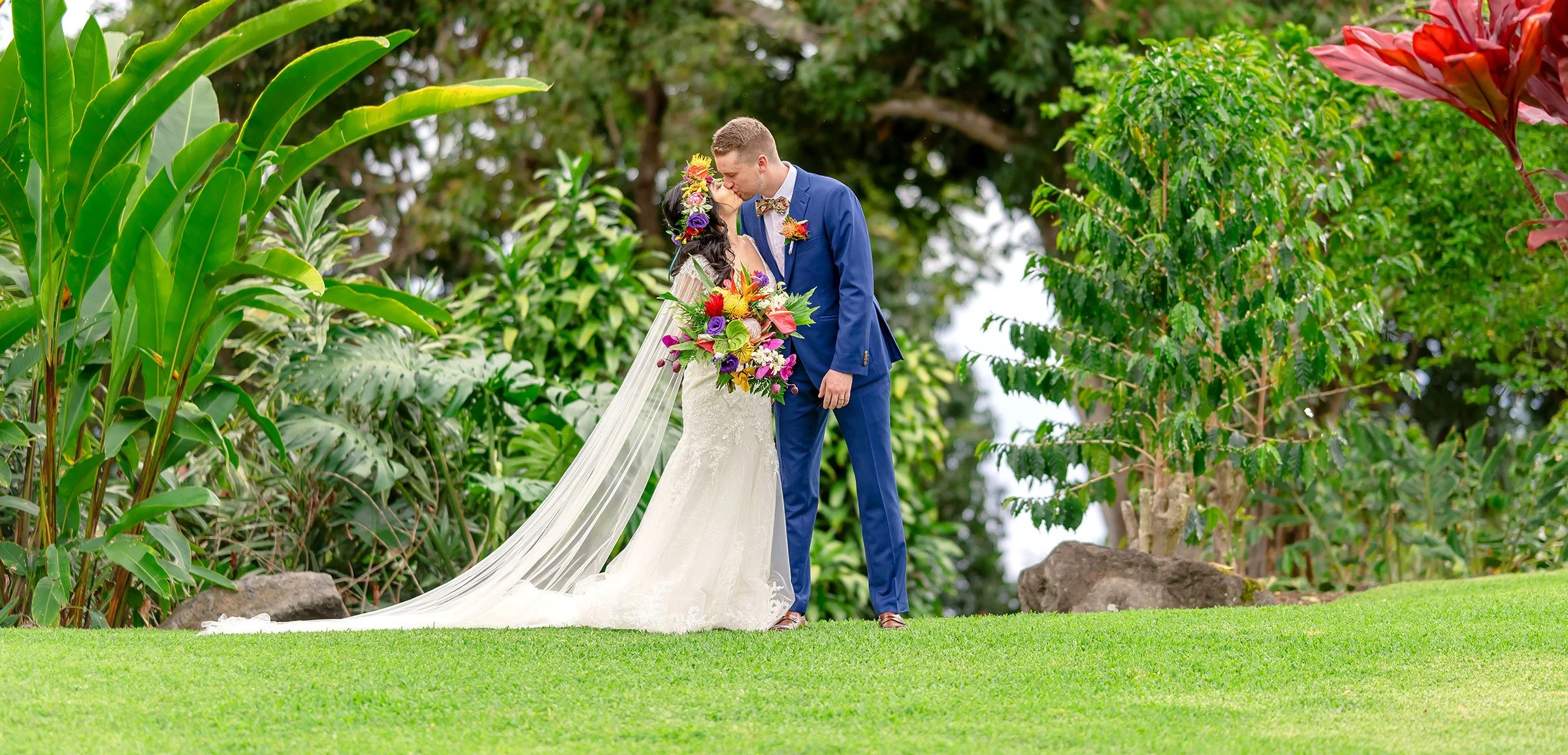 A bride in a white gown and veil holding a colorful bouquet, embraces a groom in a blue suit against a lush garden backdrop.