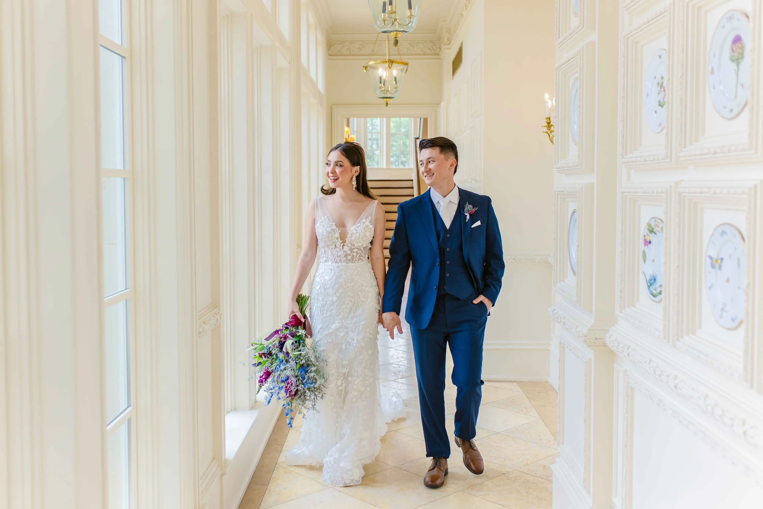 Bride and groom walking in elegant hallway, bride in lace gown with bouquet, groom in blue suit.