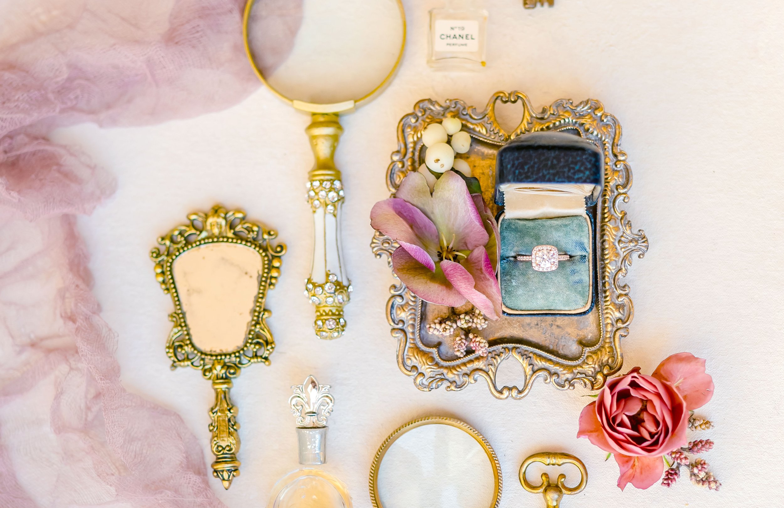 Vintage vanity items, including a gold ornate hand mirror, magnifying glass, and ring box with a diamond ring, placed on an antique tray with flowers and a perfume bottle.