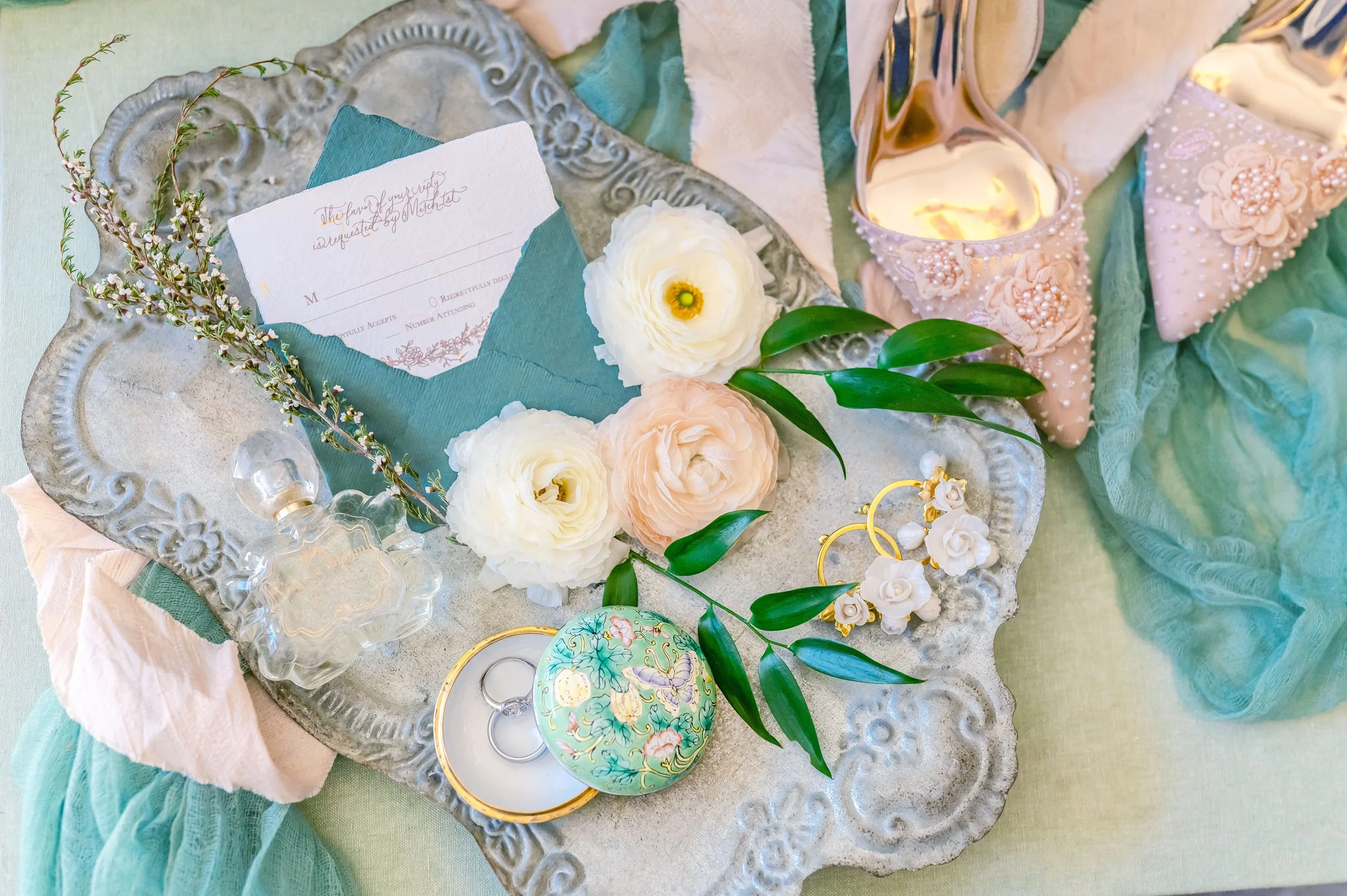 A decorative silver tray with a wedding invitation, white and peach ranunculus flowers, green leaves, a glass perfume bottle, a floral ring box with rings, gold earrings with white roses, and embellished shoes on fabric.