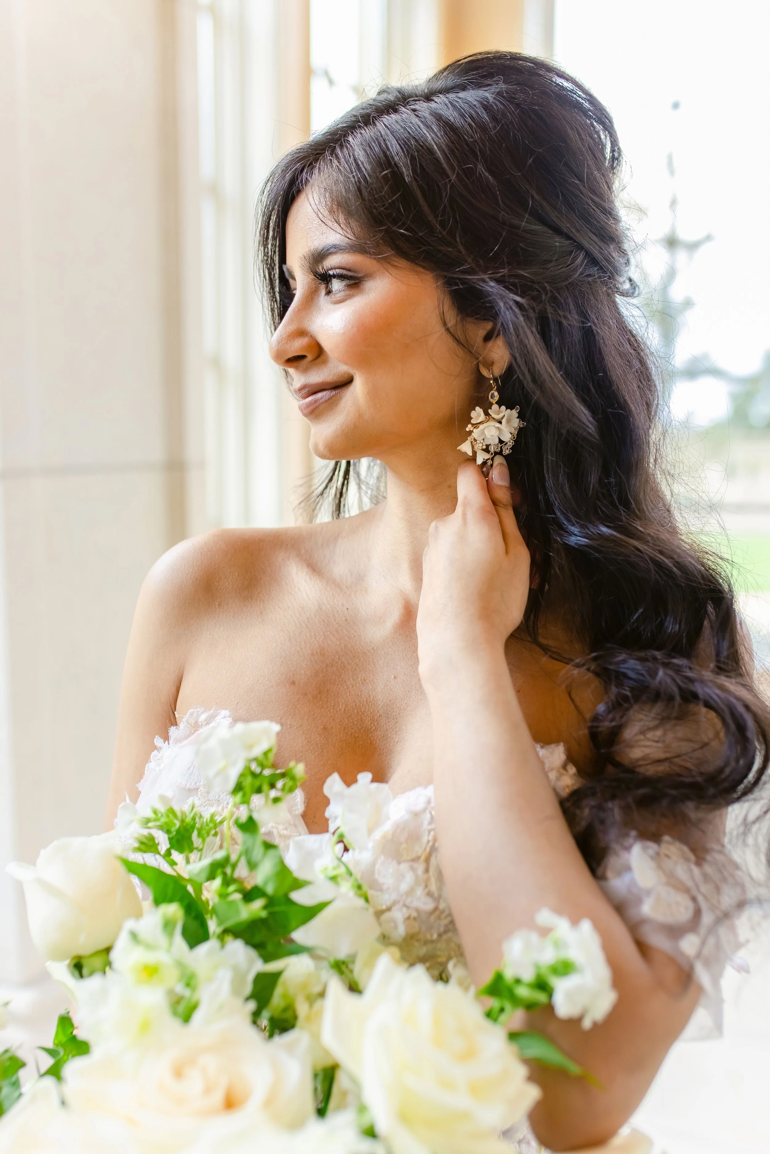 Bride holding a bouquet of white flowers, looking to the side, wearing statement earrings and a strapless wedding gown.