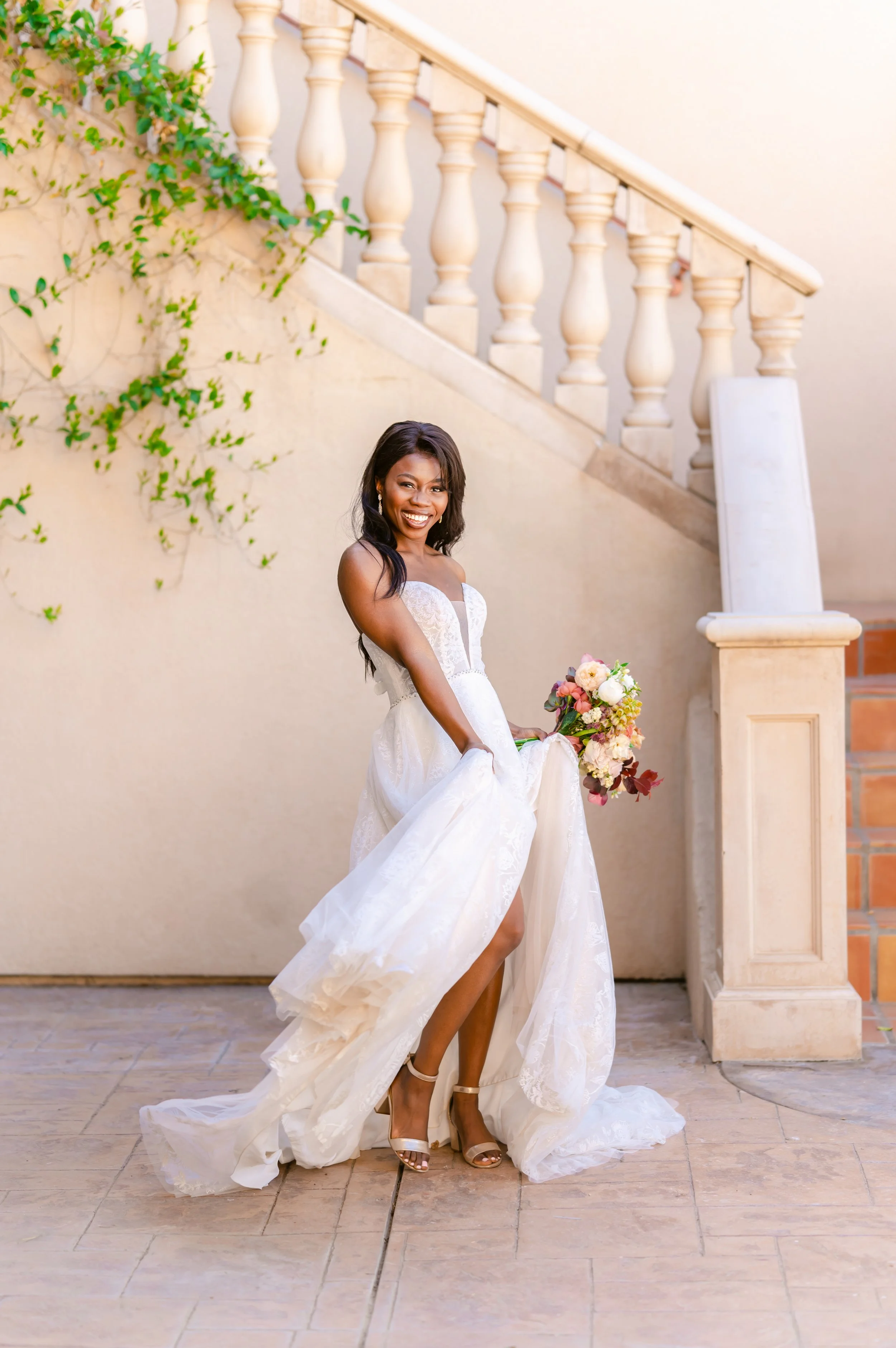 Bride in white wedding dress holding a bouquet, standing beside a staircase with greenery.
