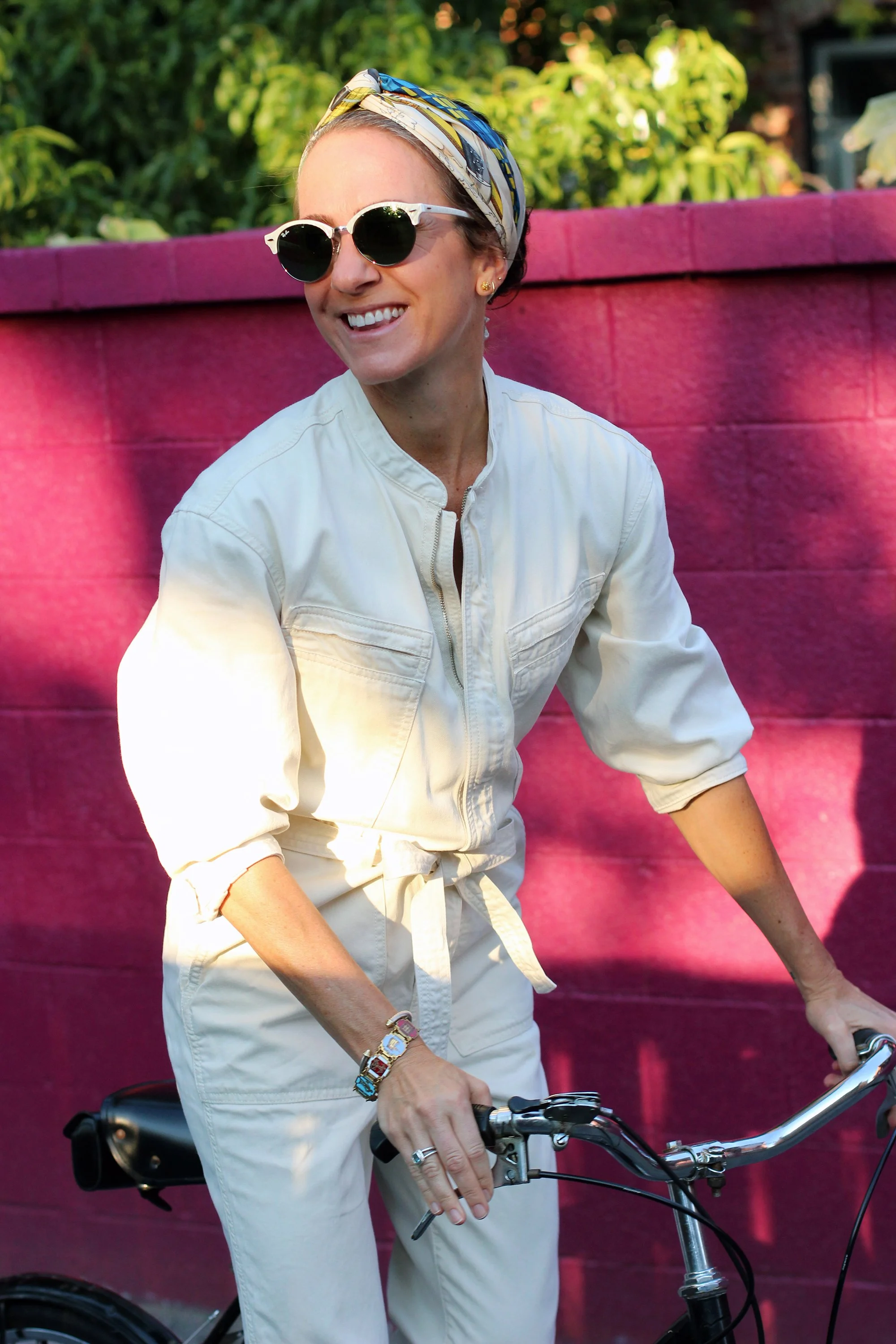 A woman wearing sunglasses, a colorful headscarf, and a light-colored jumpsuit riding a bicycle outdoors near a pink fence with green foliage in the background.