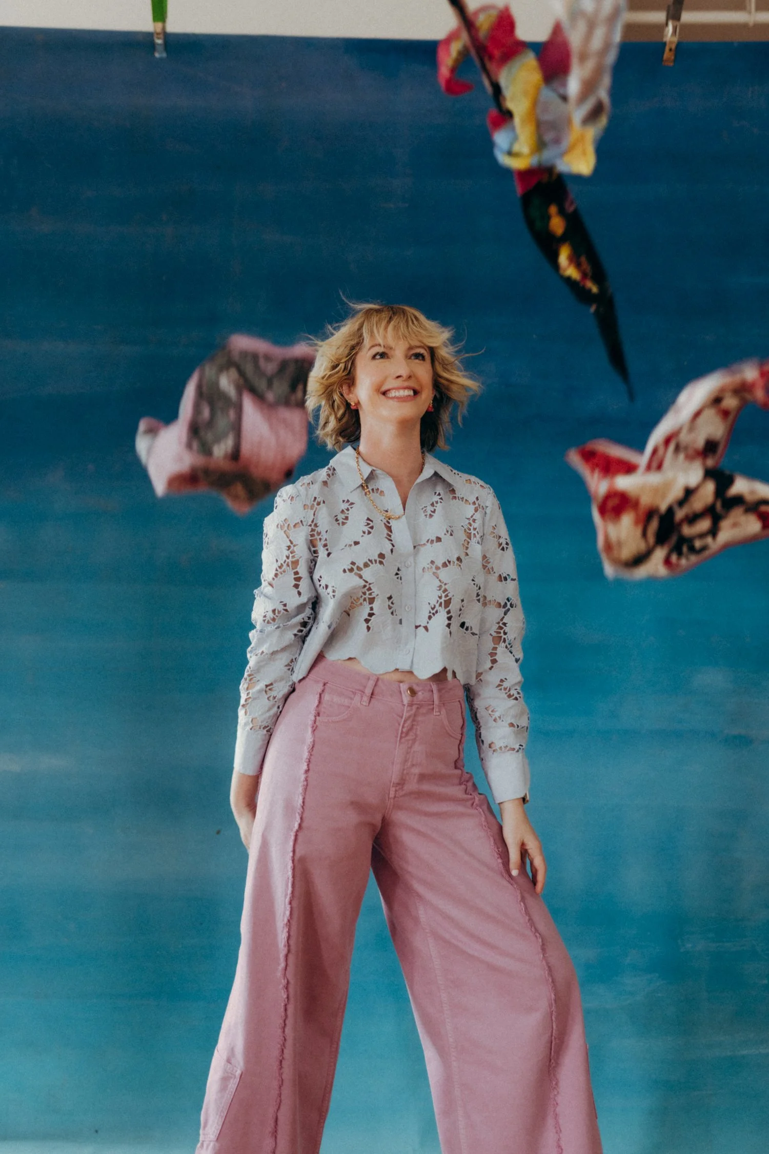 A woman with blonde hair and a cheerful expression, wearing a white lacy blouse and pink jeans, standing against a blue background with colorful fabric pieces hanging above her.