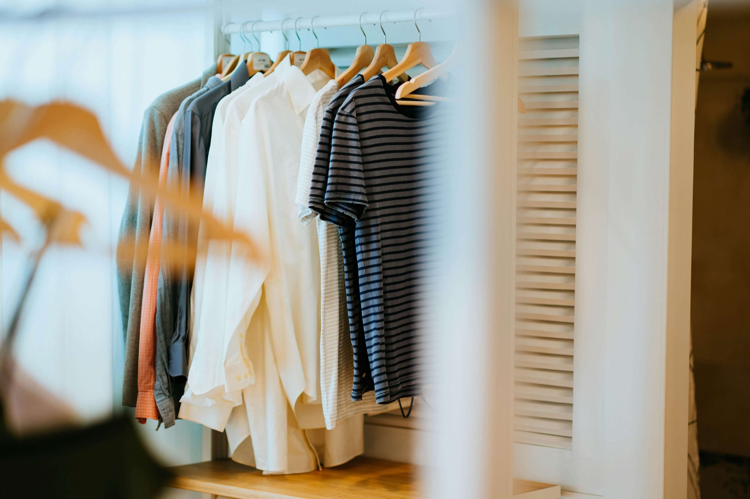 A closet with a hanging rail of men's clothes including shirts and t-shirts, with a wooden louvered door on the right side.