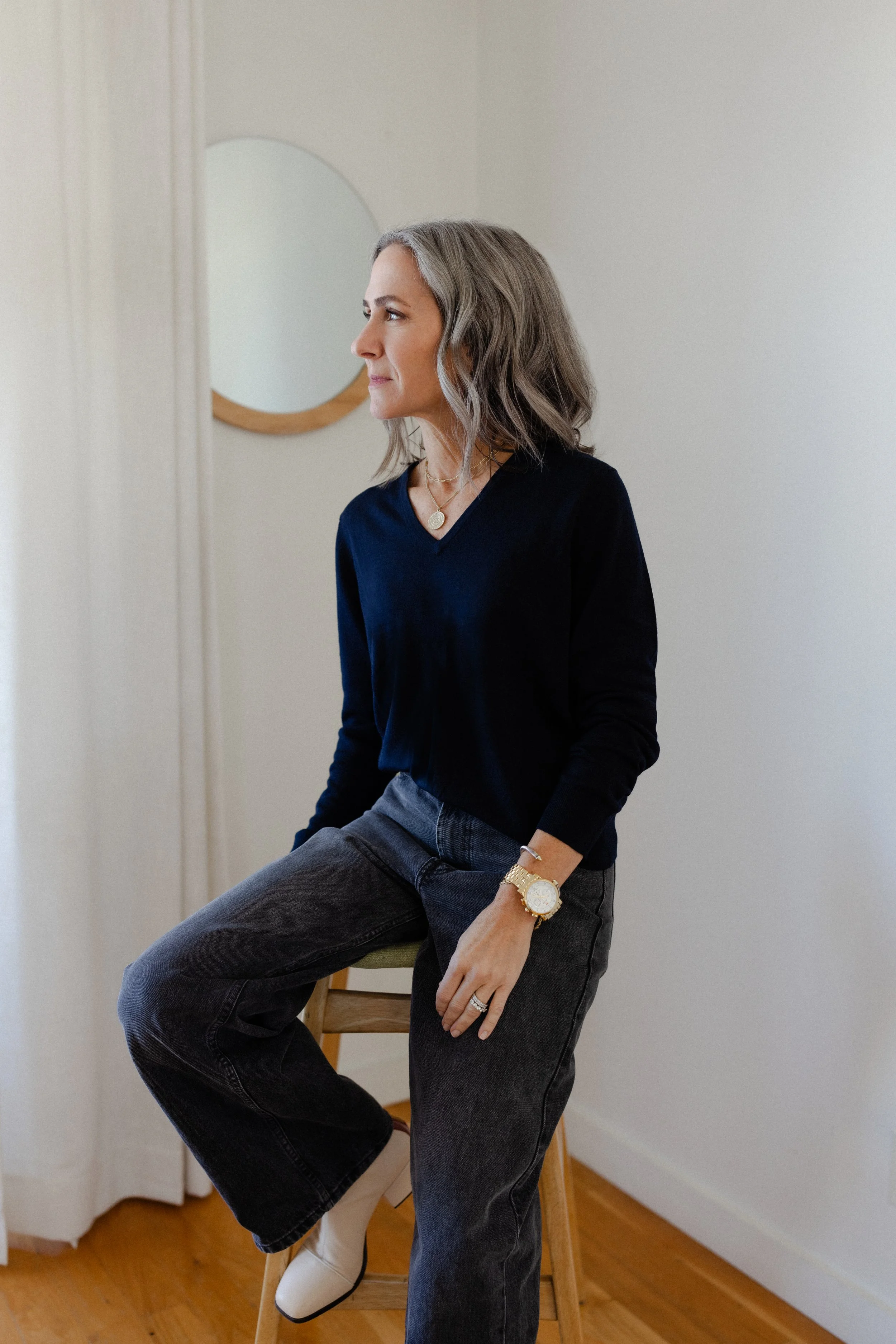 A woman with gray shoulder-length hair wearing a dark blue V-neck sweater, gray pants, white ankle boots, gold jewelry, and a gold watch, sitting on a wooden stool in a room with hardwood floors and white walls, looking to her left.