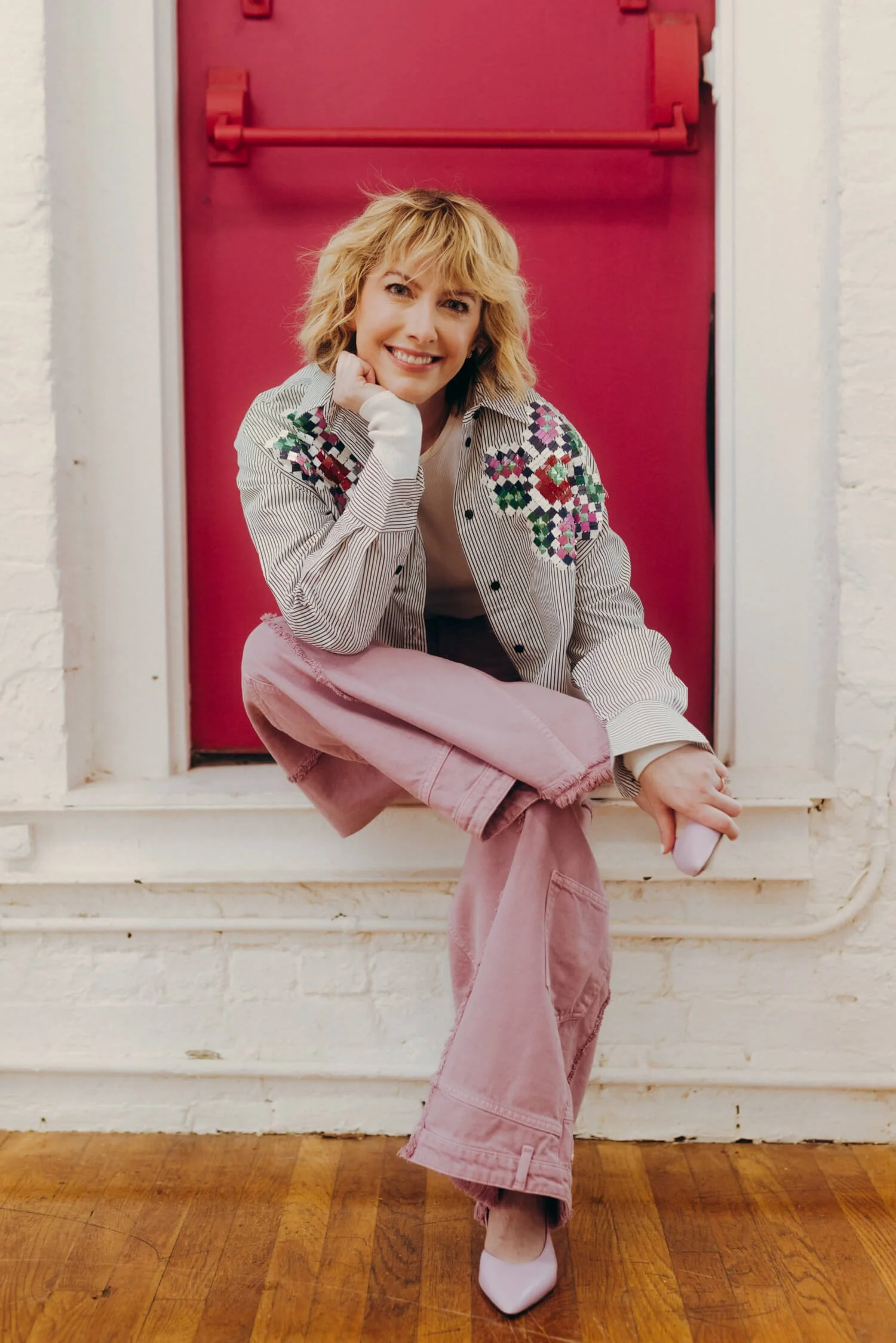 A woman with short blonde hair and a bright smile, wearing a plaid shirt with colorful embroidery on the shoulders, pink pants, and white boots, sitting on a window ledge in front of a red door with a horizontal bar.