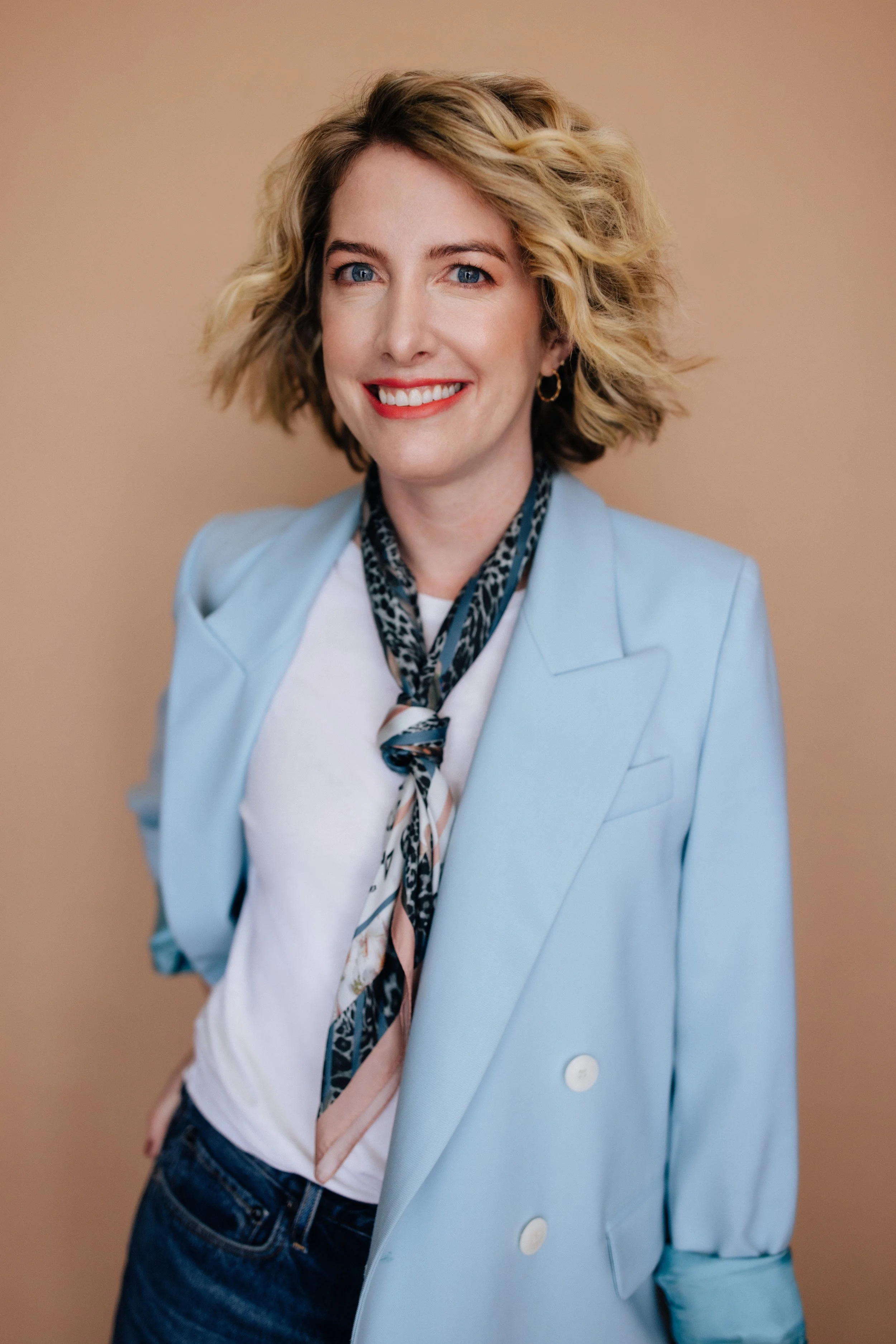 A woman with blonde, curly, shoulder-length hair and blue eyes, smiling with red lipstick, wearing a light blue blazer, a white shirt, and a patterned scarf, standing against a beige background.