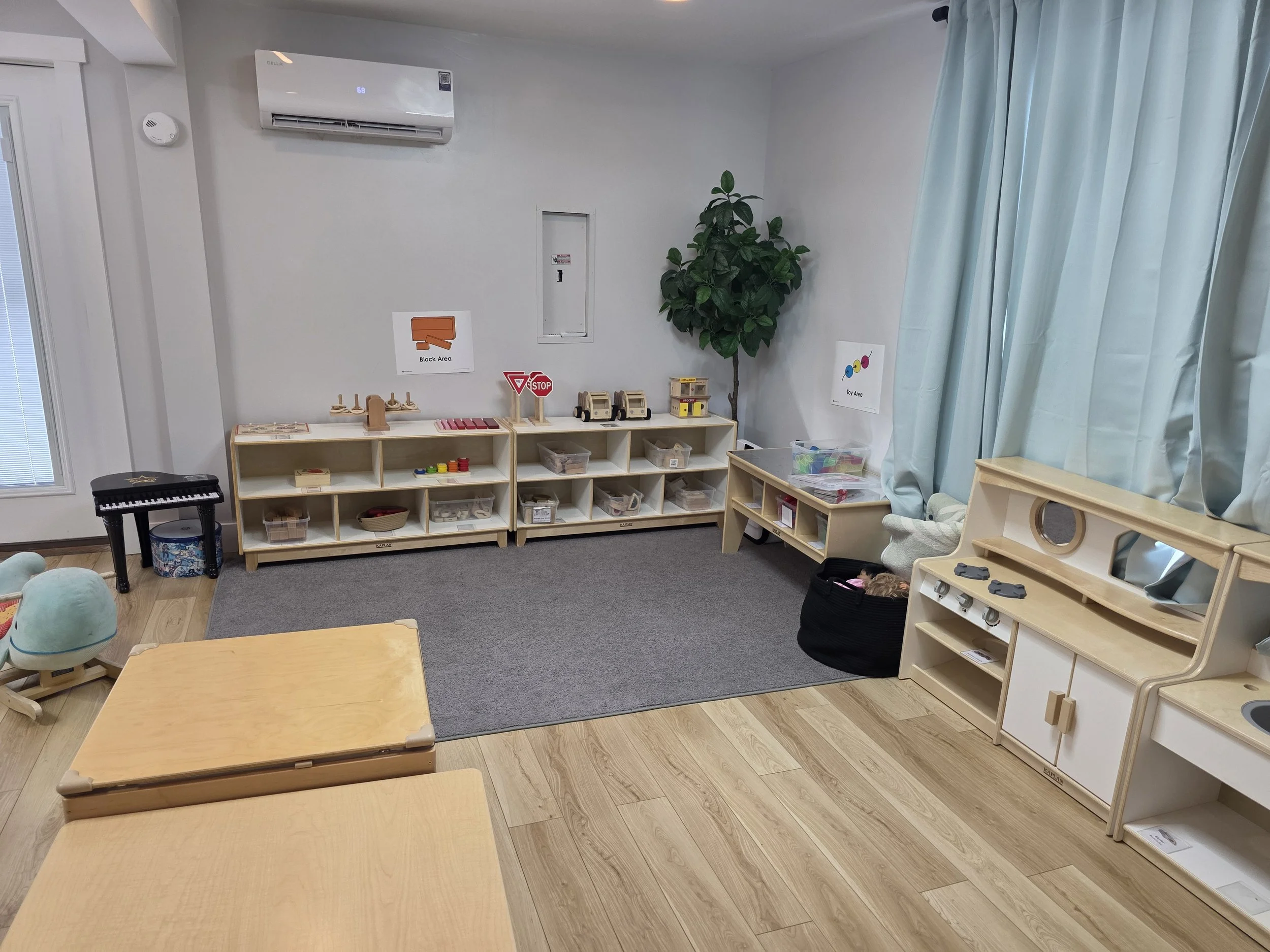 A children's play area with wooden shelves filled with toys, a small piano, and some stuffed animals. There is a green leafy plant, window with blinds, light blue curtains, and signs indicating areas for blocks and toys.