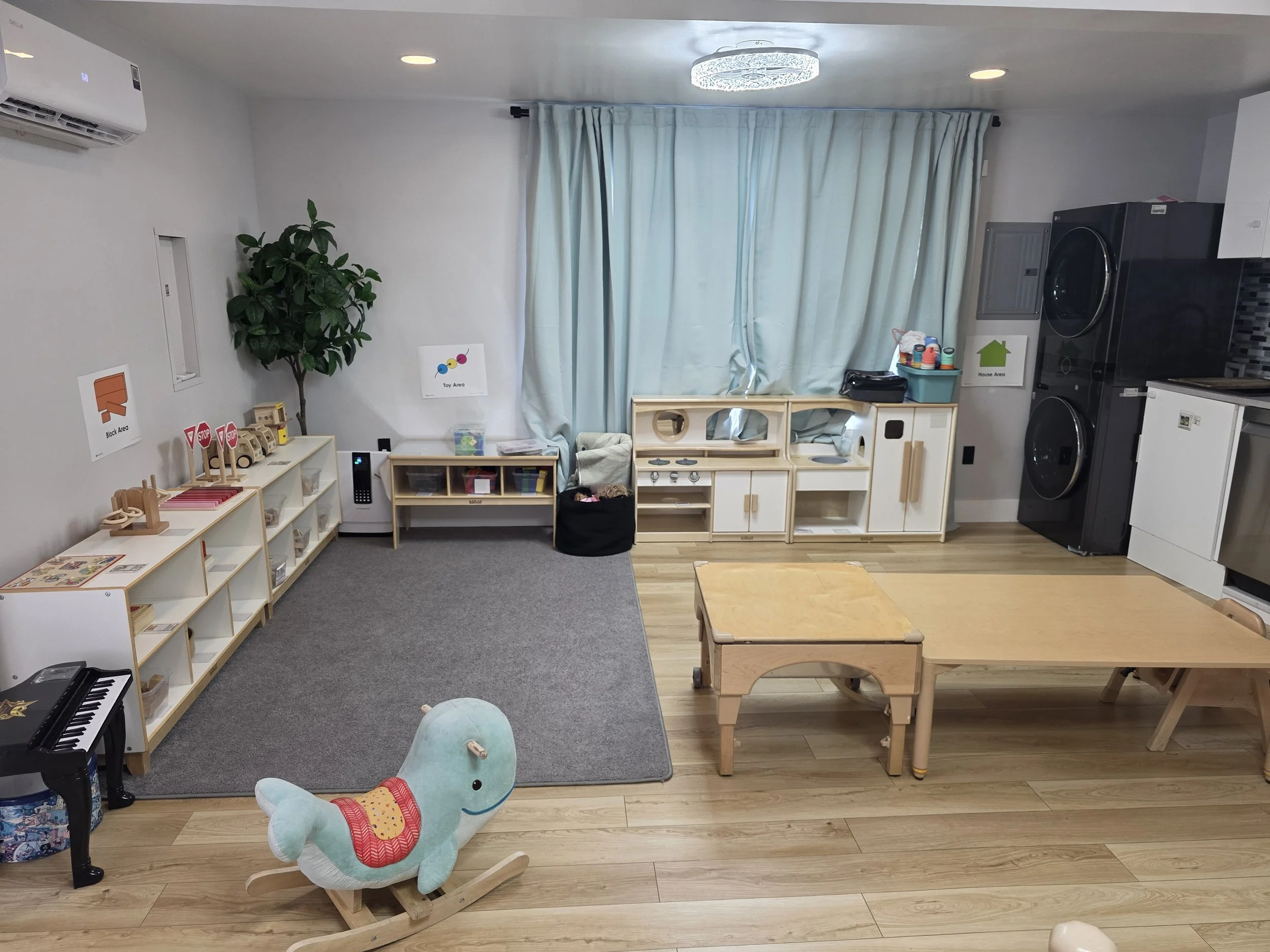 A children's playroom with a wooden rocking horse in the center, a small table and chairs, storage units labeled for toy and house areas, a potted plant, plush toys, and laundry appliances in the background.