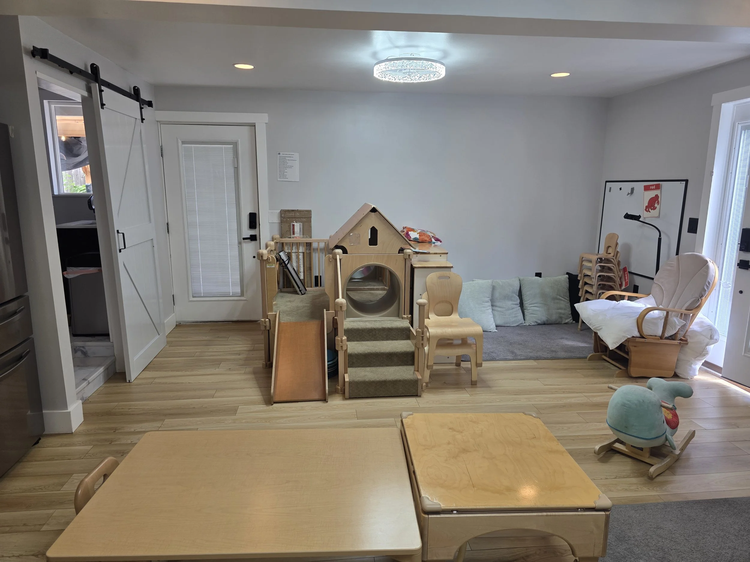 A room with light wooden flooring and white walls, featuring children's furniture including a small slide, a dollhouse, a rocking chair, and a stack of wooden chairs. There is a white door with vertical blinds, a whiteboard with a red illustration of