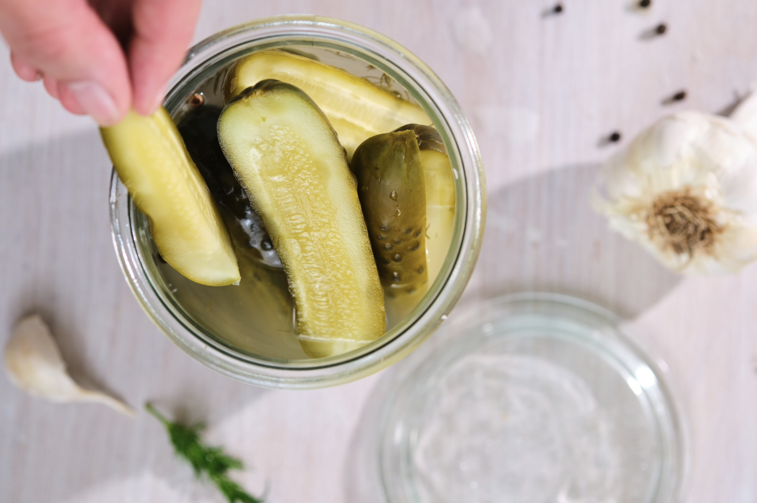 A glass jar filled with pickles and cucumber slices submerged in liquid, with a garlic bulb and a sprig of dill nearby on a light wooden surface.