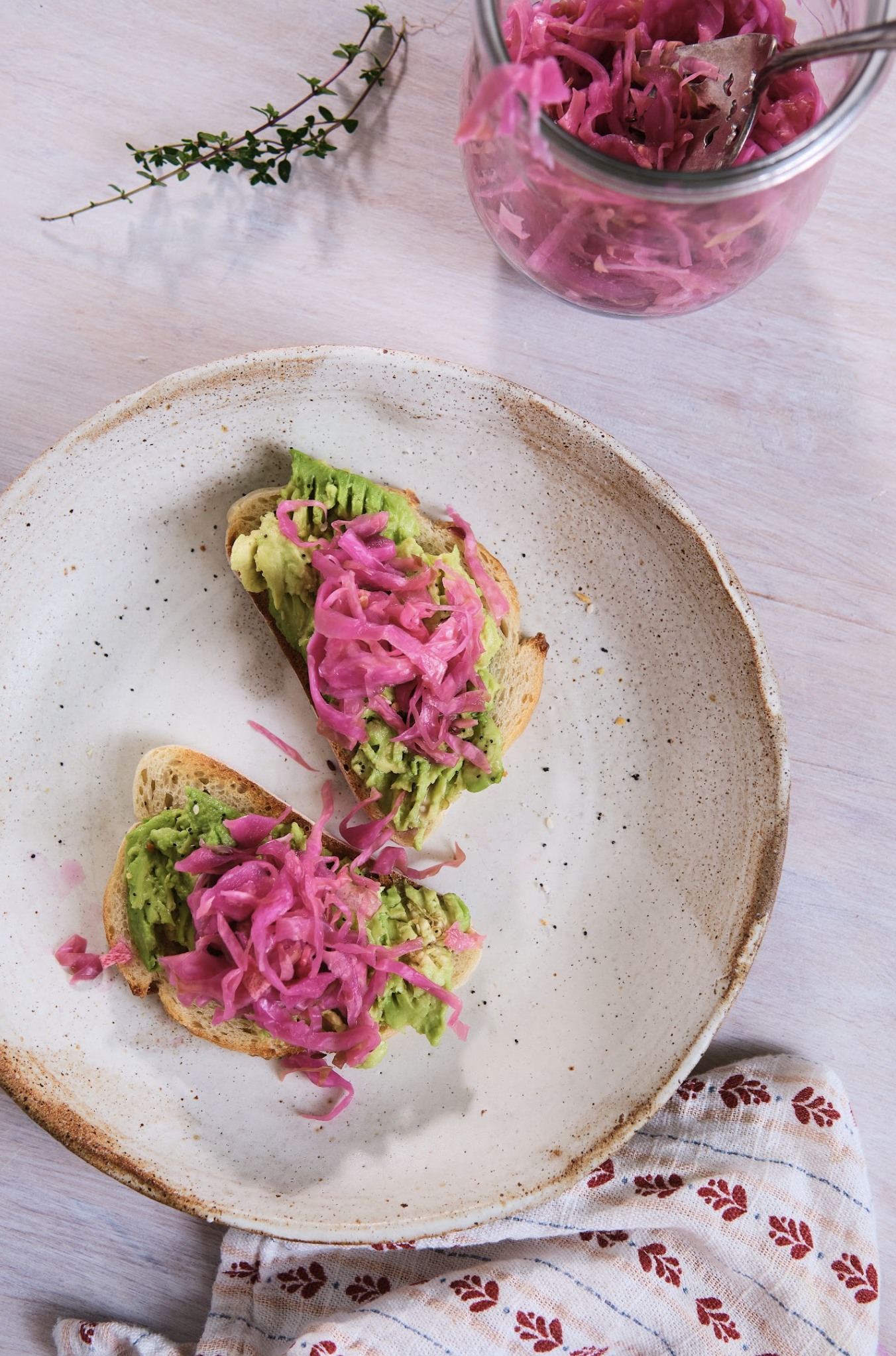 Open-faced sandwiches with mashed avocado and pickled red onions on a ceramic plate, placed on a table with a cloth napkin that has a red leaf pattern. A small jar of pickled red cabbage is visible in the background, along with a sprig of fresh herb.