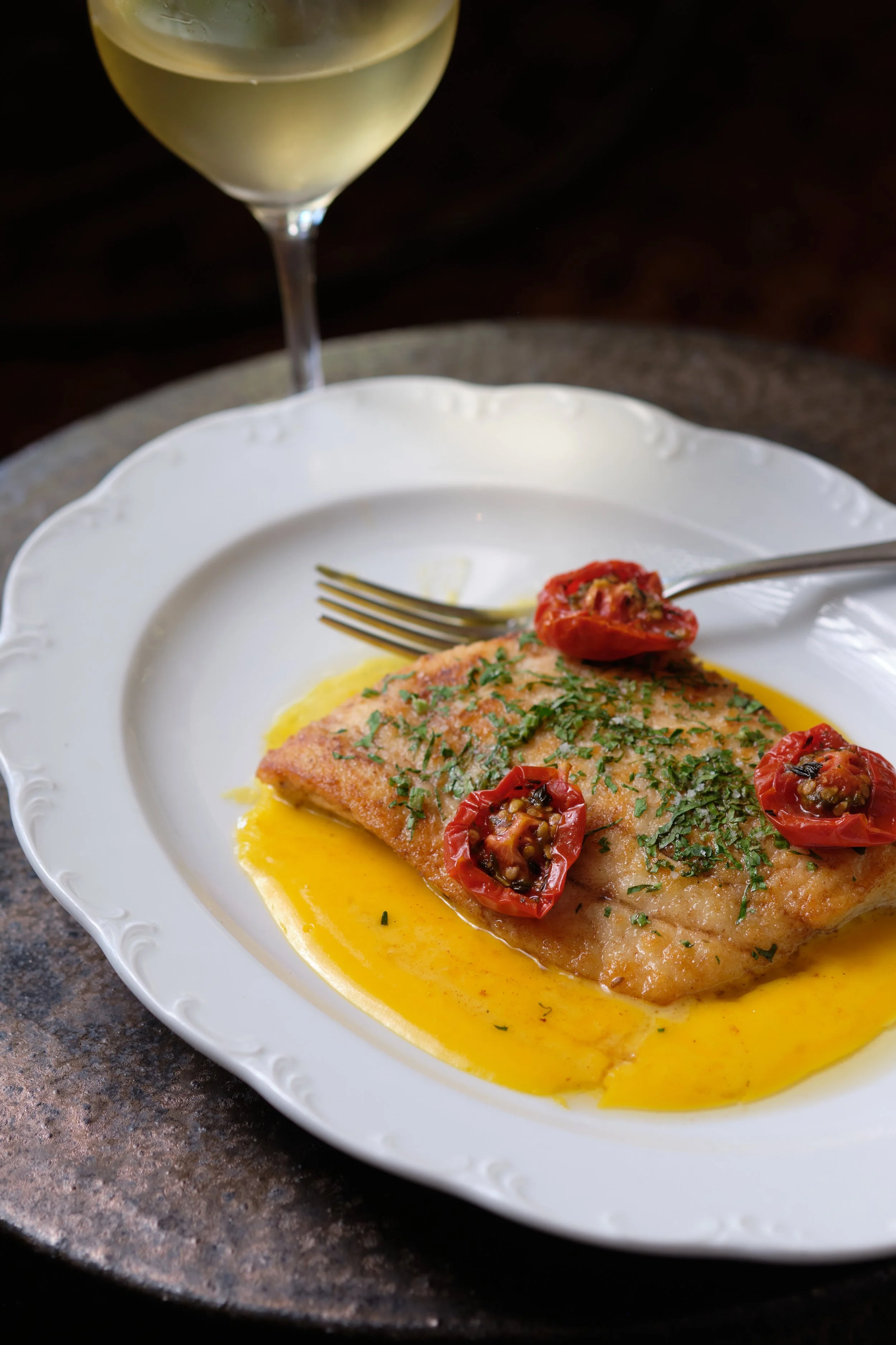 A white plate with a cooked fish fillet topped with cherry tomatoes and chopped herbs, drizzled with lemon butter sauce. A fork rests on the plate, and a glass of white wine is in the background.