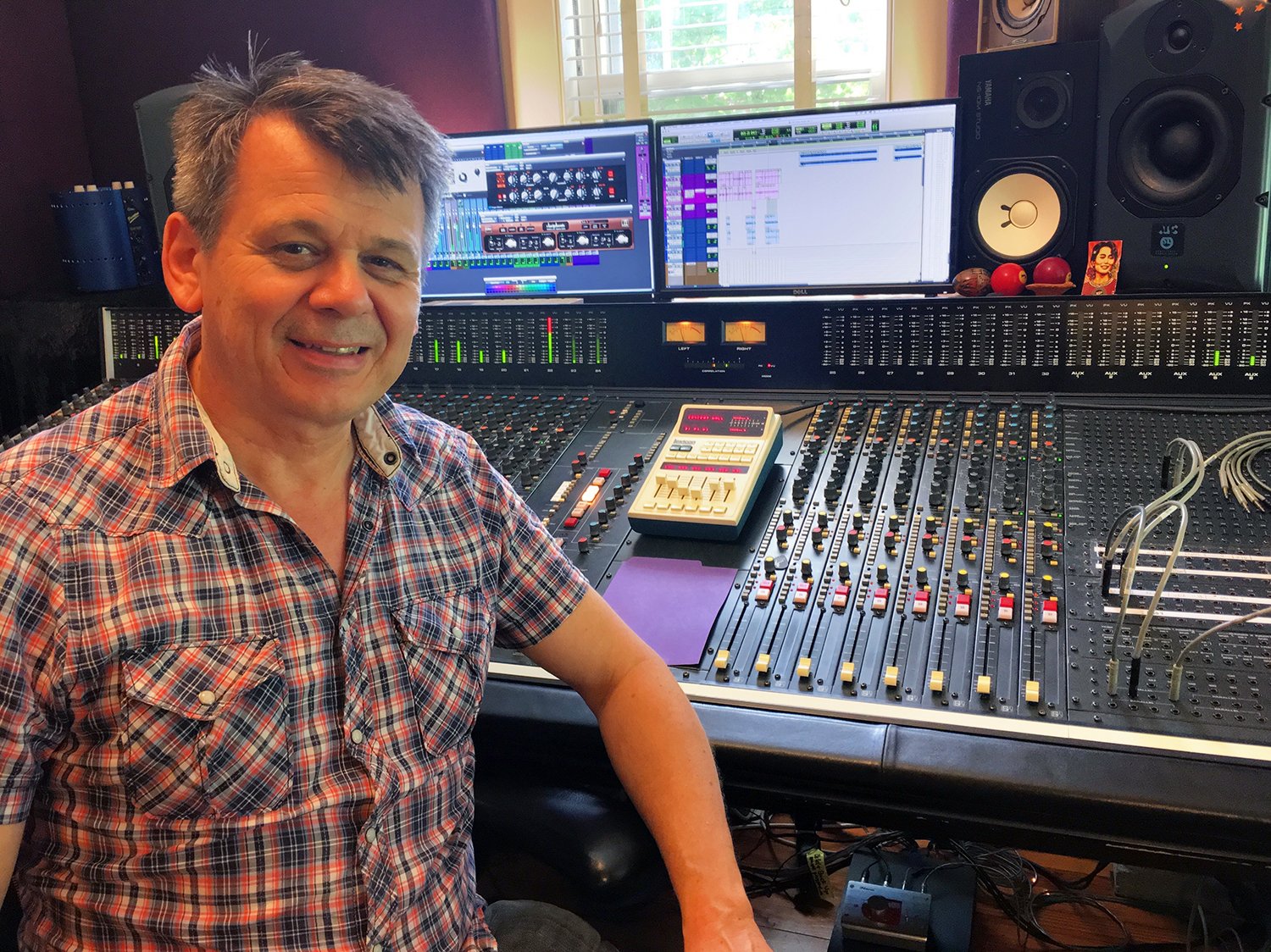 Pal Sheldon, seated in a recording studio with a large mixing console, computer screens displaying audio editing software, studio monitors, and various audio equipment.