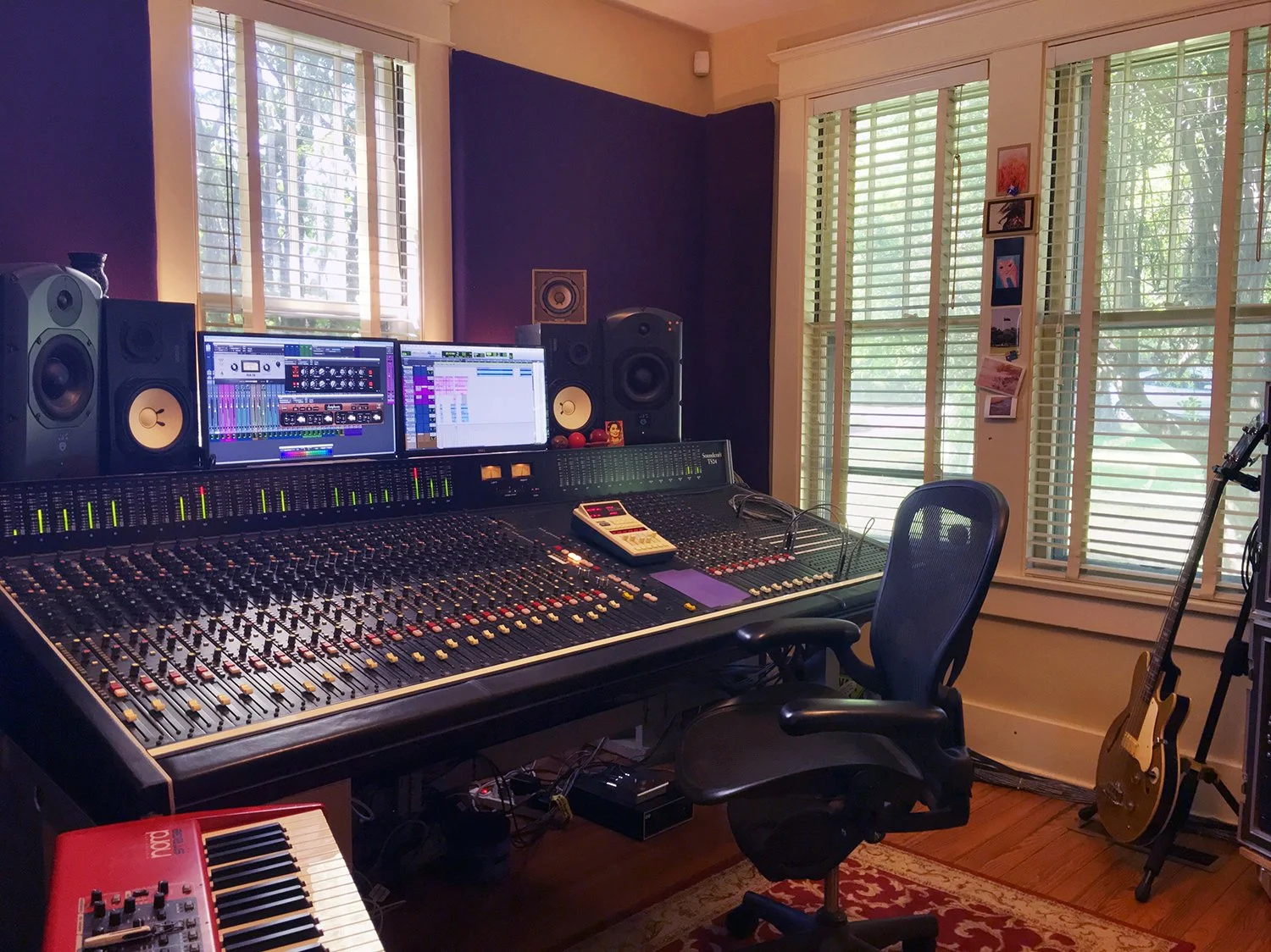 A music studio with a large mixing console, two computer monitors displaying audio editing software, multiple speakers, a keyboard, and a guitar on a stand near the window.