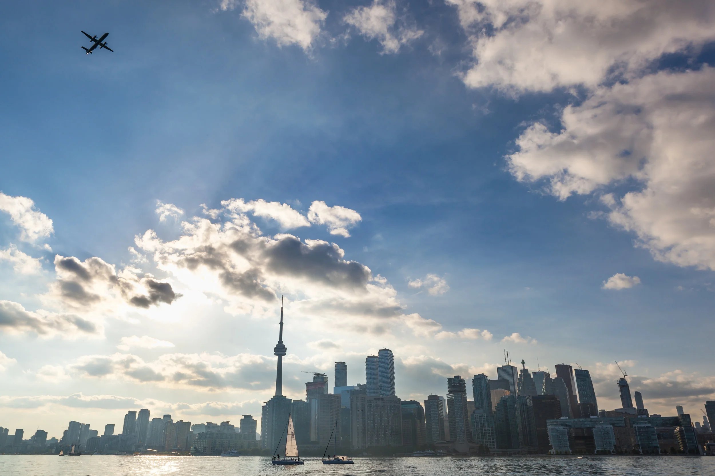 Toronto daytime cityscape; sail boats, majestic clouds, and plane above.