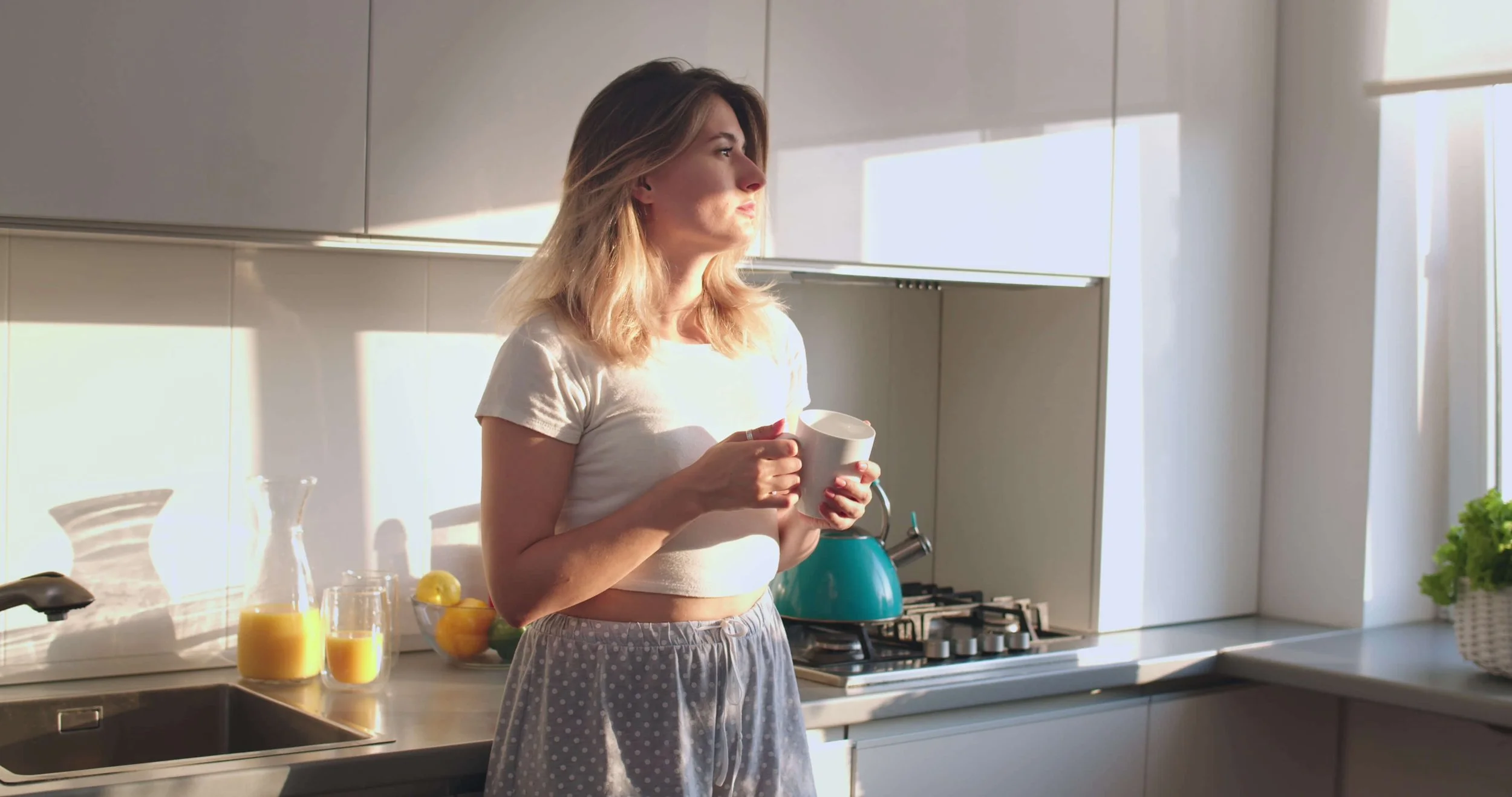 Woman standing in kitchen holding a coffee mug, looking tired and reflective, illustrating why women’s energy and metabolism change even when nothing in their diet or lifestyle has shifted.
