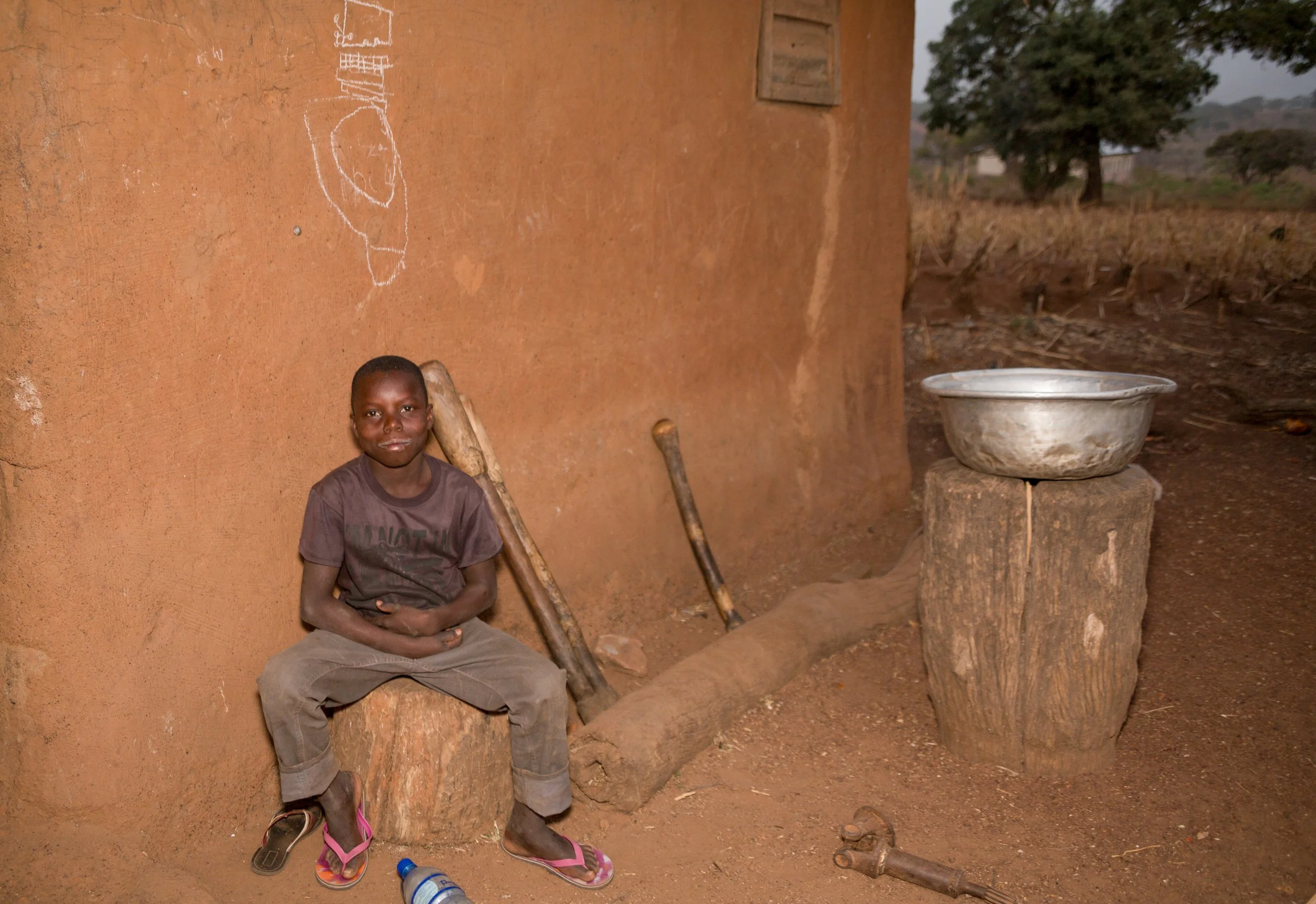 Assis contre le mur rouge de la maison en terre, Lucien se repose. L’école perdue demeure en arrière-plan.
