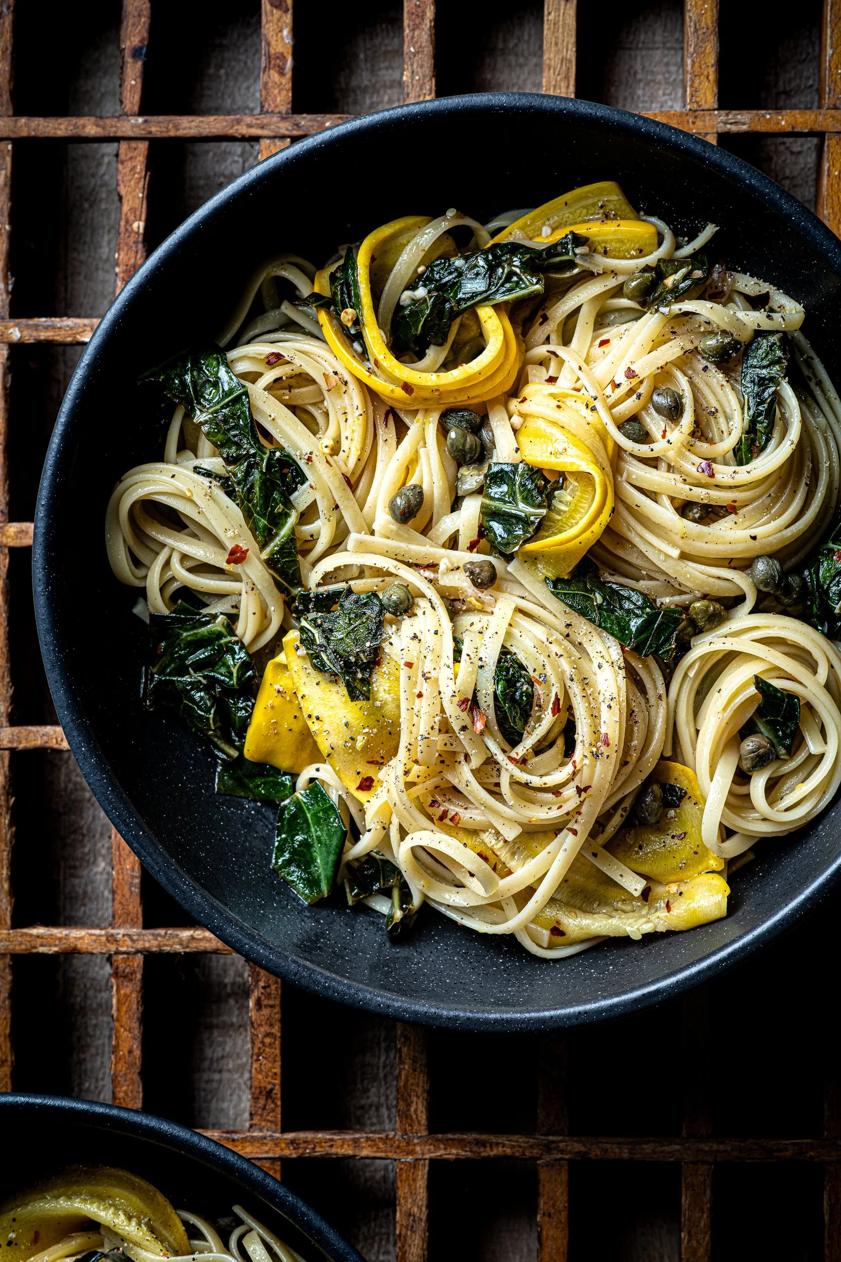 A bowl of linguini pasta with ribbons of zucchini, sauted kale and capers