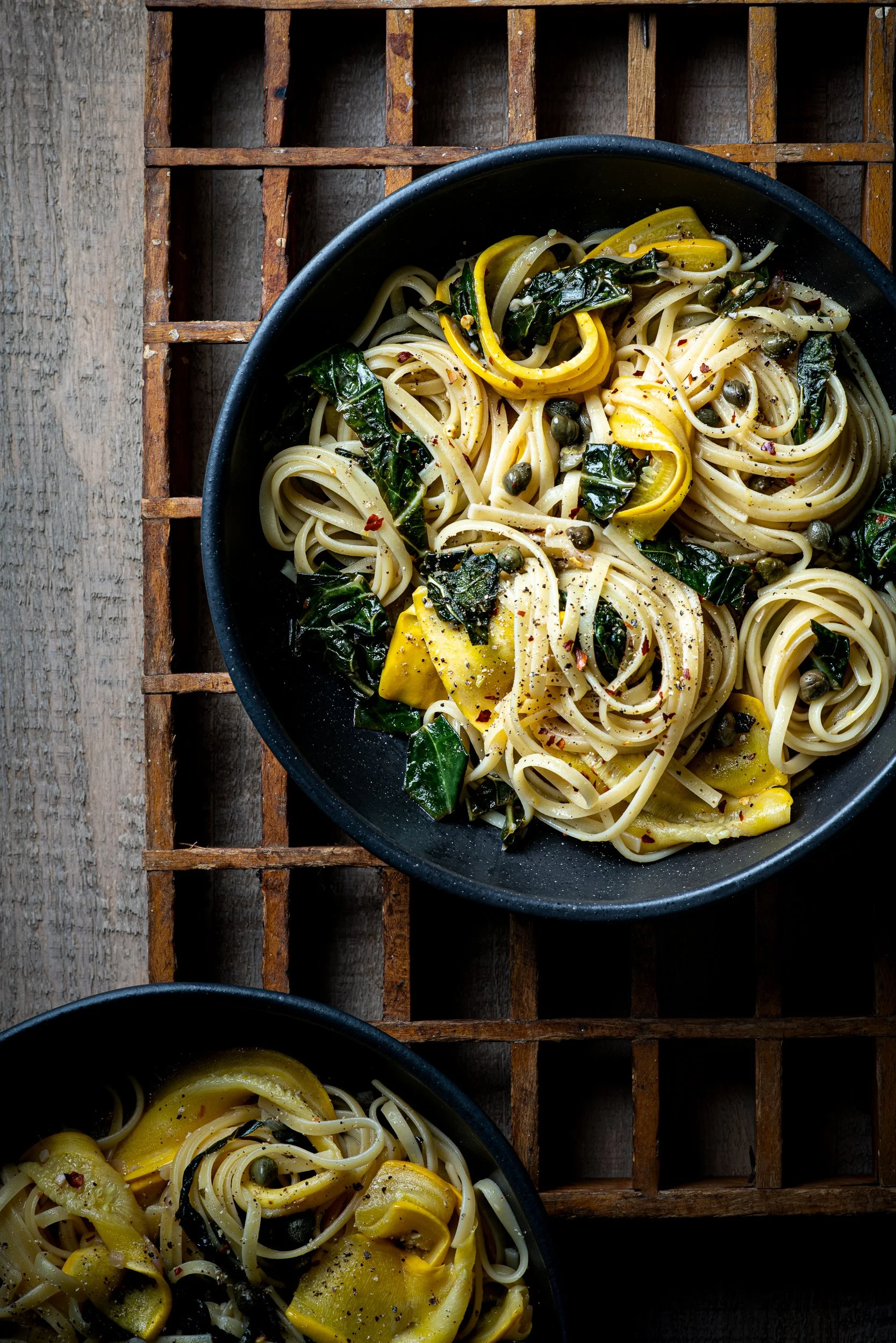 A bowl of linguini pasta with ribbons of zucchini, sauted kale and capers