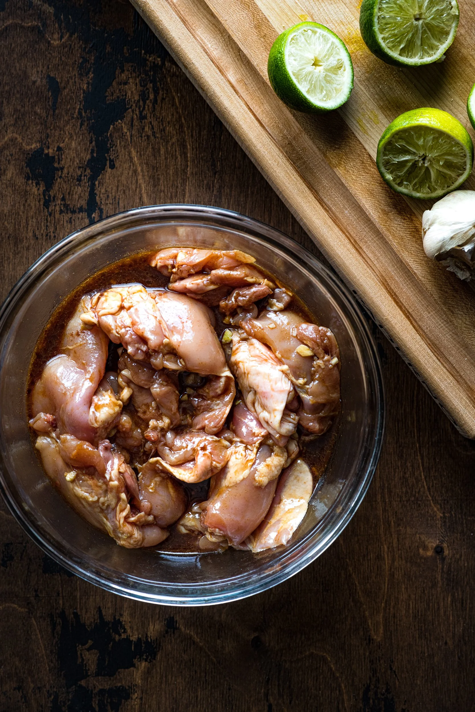 Raw chicken in a bowl tossed in marinate next to a cutting board with limes cut in half