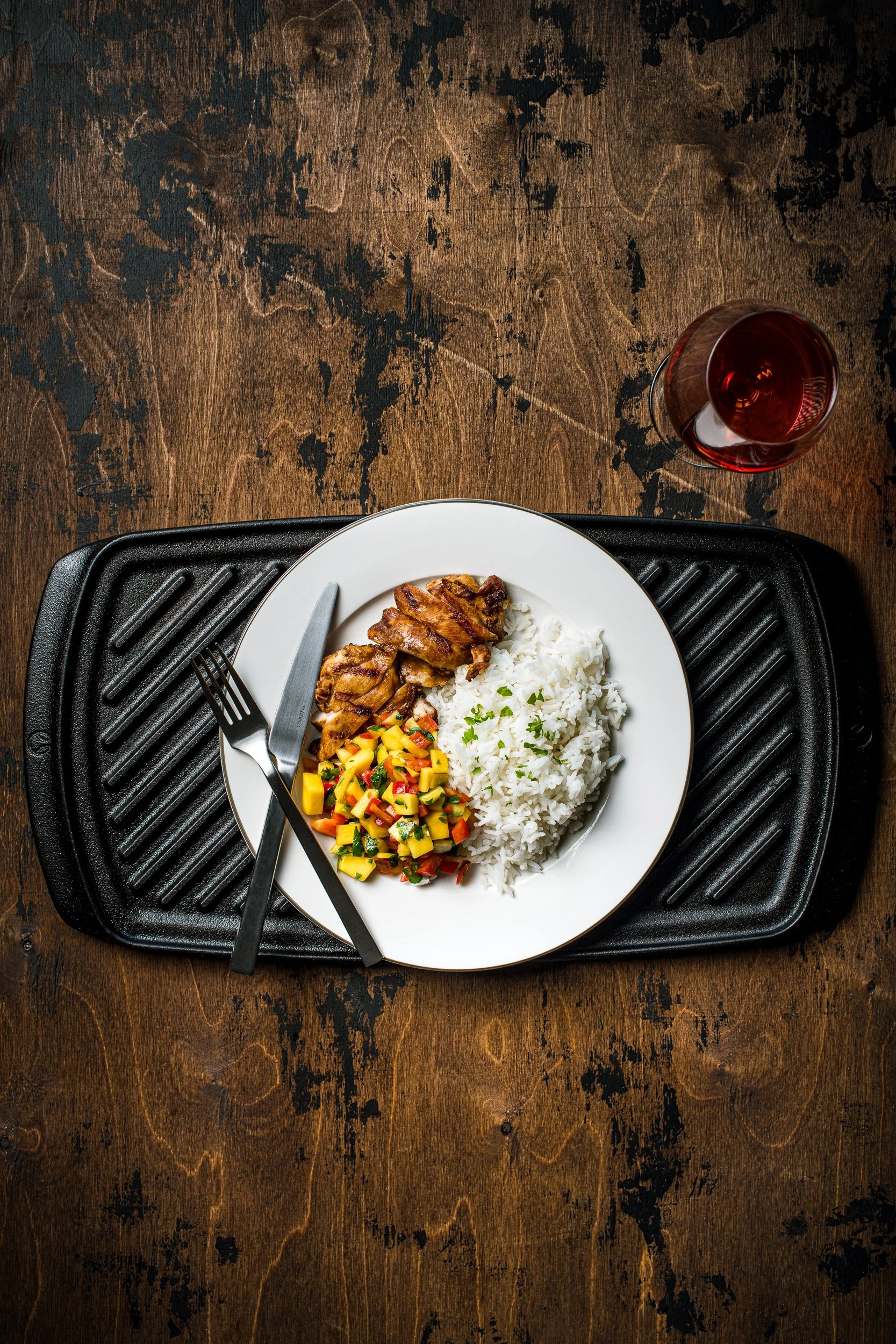A plate of grill chicken, white rice and mango salsa that is on a cast iron grill pan on a wood table. Next to a glass of wine. A fork and knife is on the left side of the plate