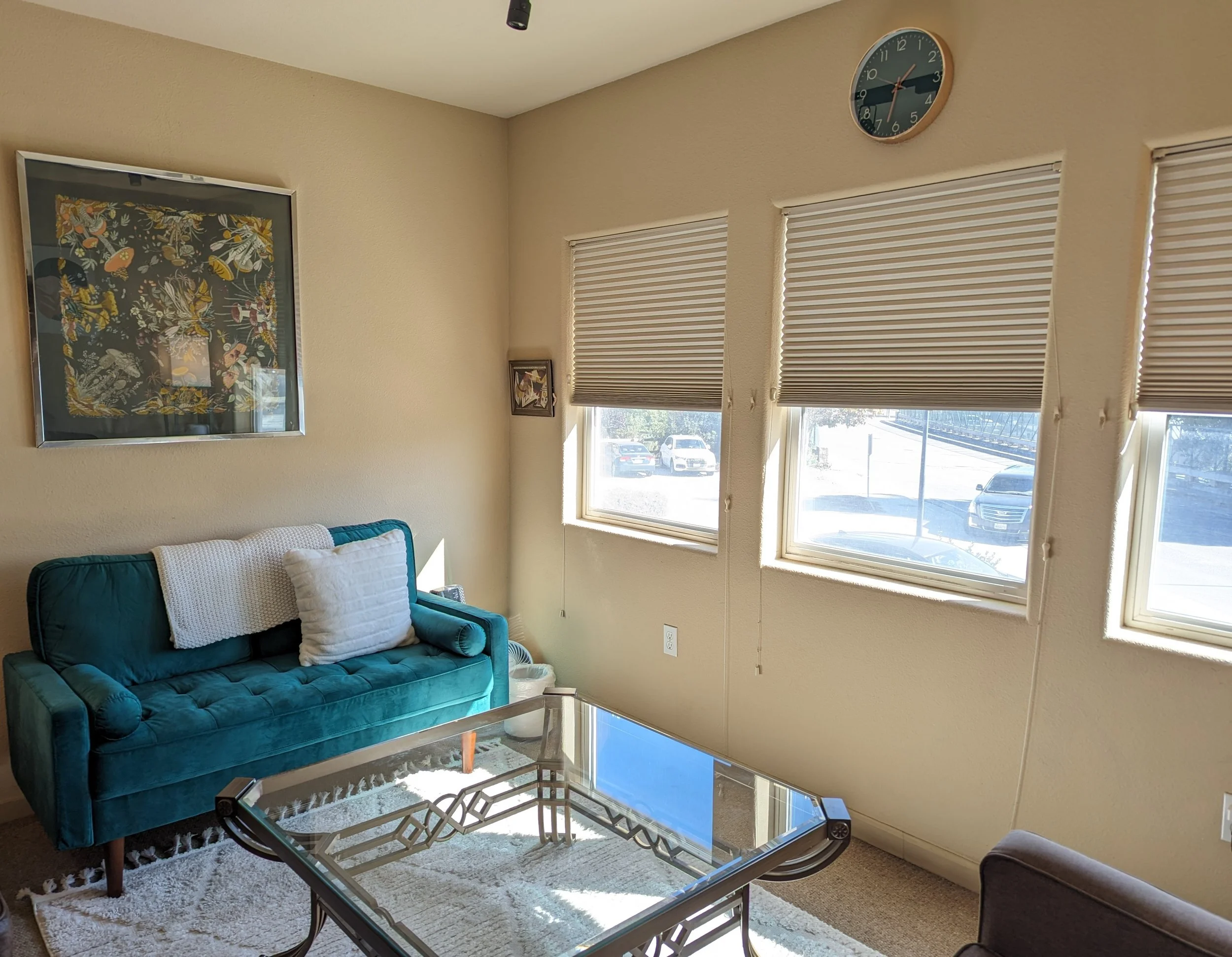 Living room with cream-colored textured walls, a teal velvet sofa with white pillows, a glass coffee table with a decorative metal frame, three windows with blind shades, and a large wall clock showing 12:12. There is a framed artwork on the wall and a small picture beside the windows.
