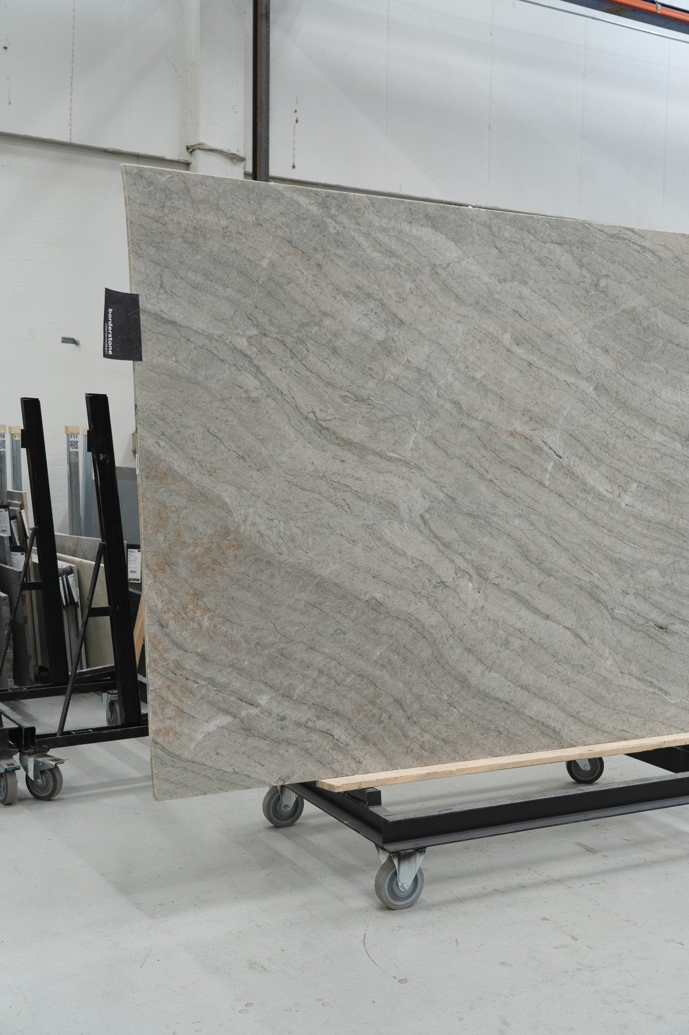 En stor plade af lysegråt natursten med blodrøde og beige nuancer, står på en vogn med hjul i et lager.