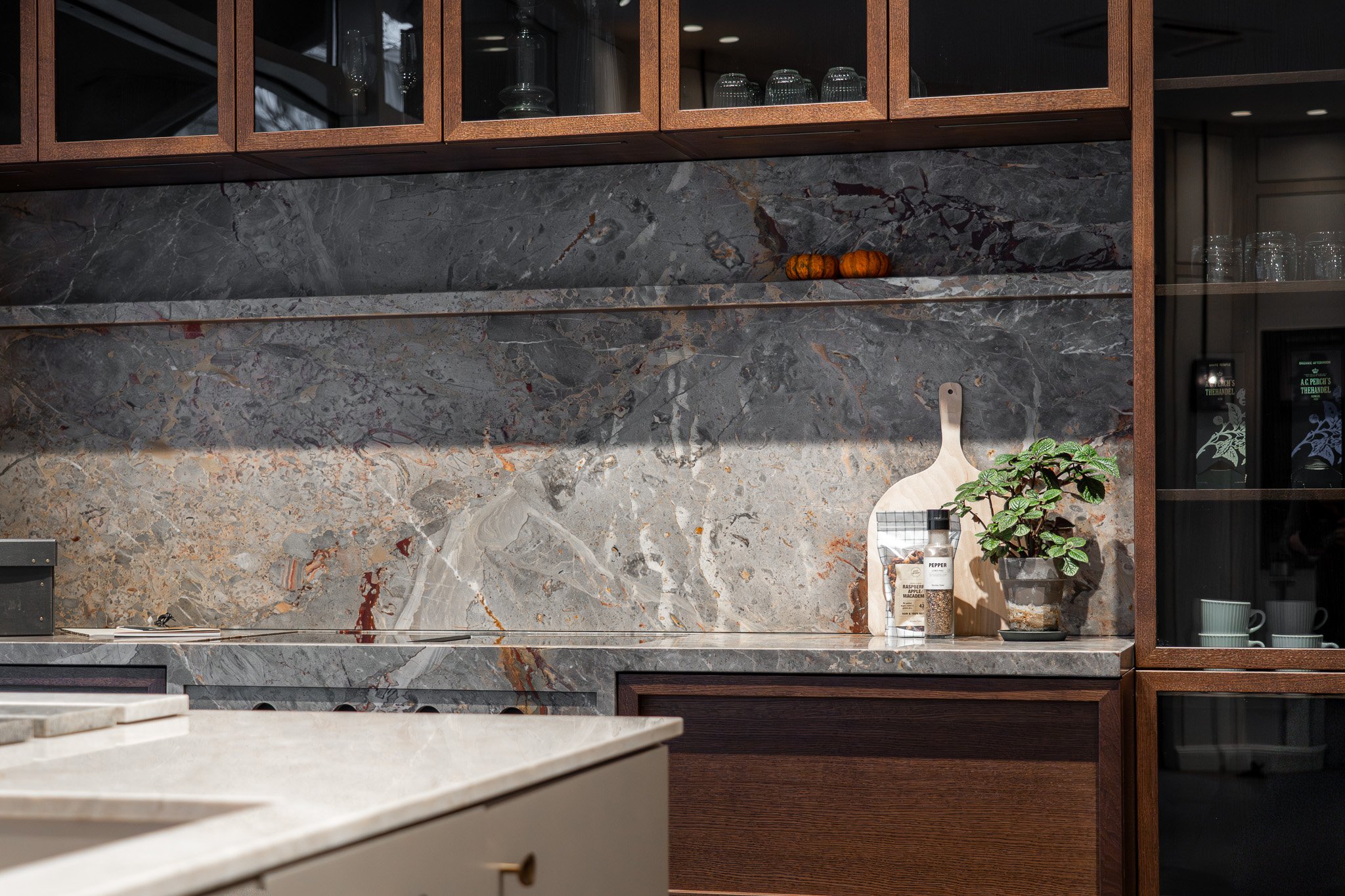 Køkken med mørkegrå marmor backsplash og træskabe. Der er en stor ø i midten med en lysbordplade. På væggen er der små dekorative gulerødder og en grøntsagskniv. Der er en grøn plante og en beholder med krydderier på bordet.