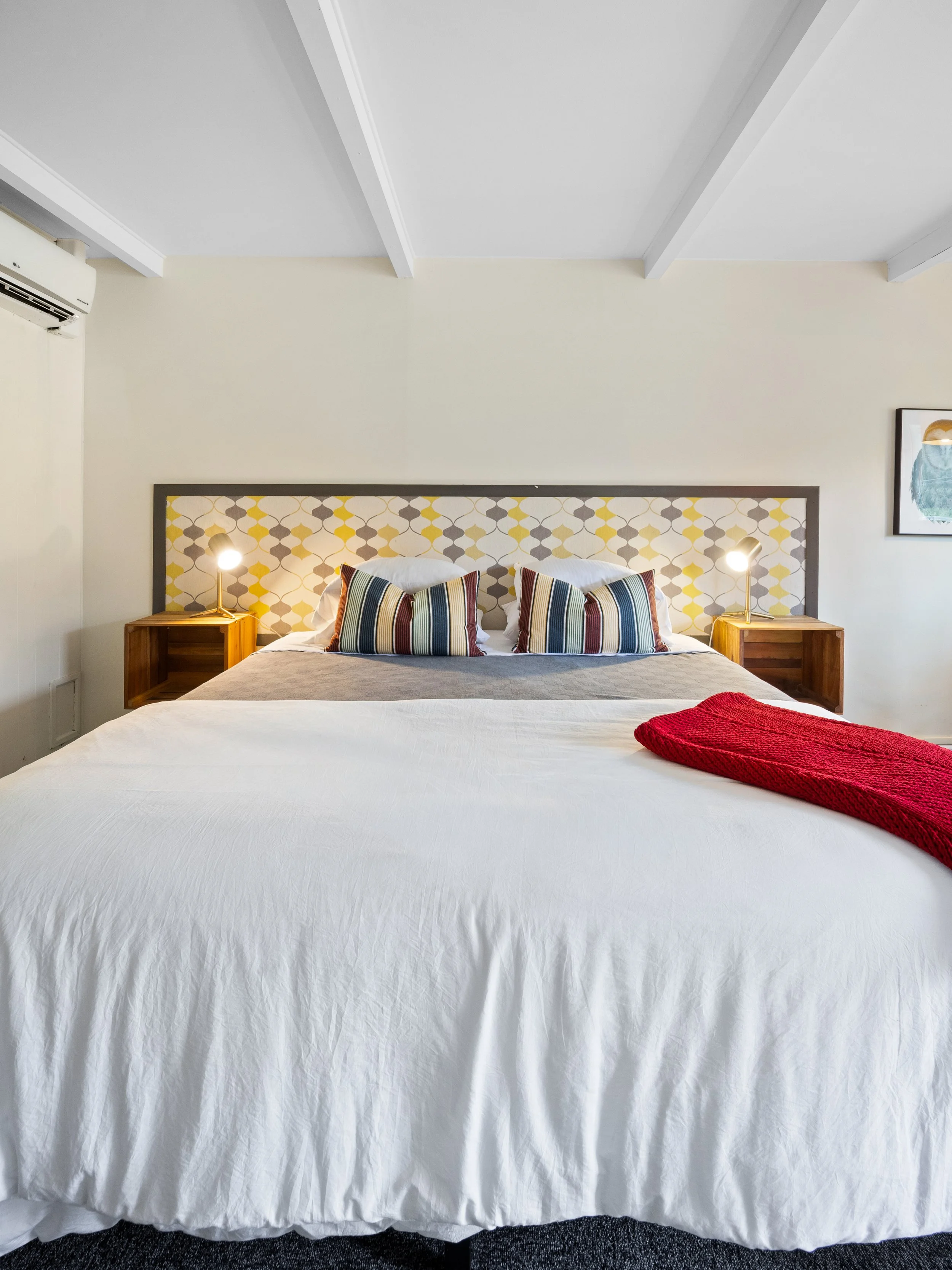 A neatly made bed with white sheets, four striped pillows, a red blanket at the foot of the bed, wooden nightstands with small lamps on each side, a patterned headboard, a framed picture of a bird on the wall, and an air conditioning unit on the wall.