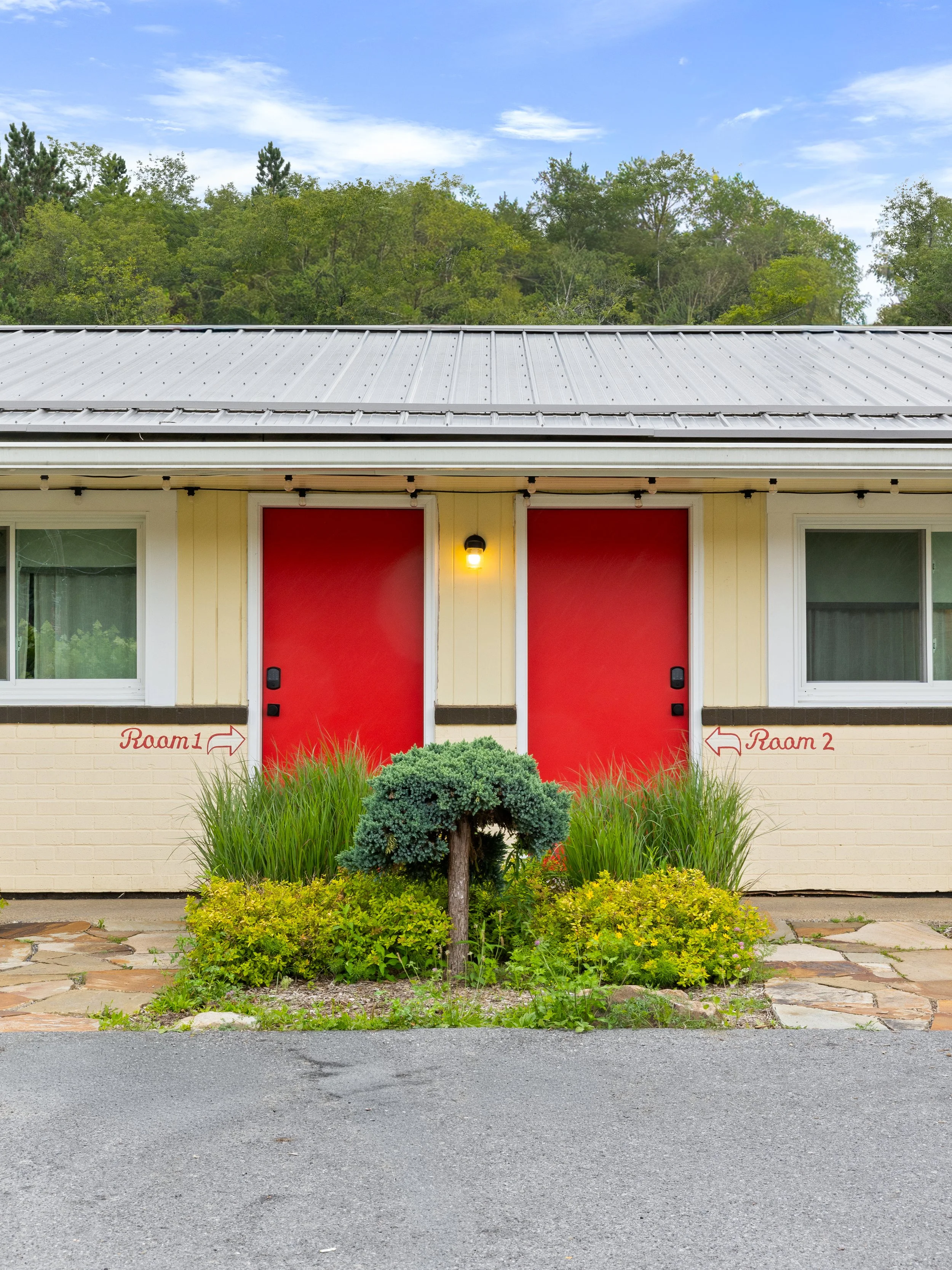 Front view of a building with red doors labeled 'Room 1' on the left and 'Room 2' on the right. There is a small landscaped garden with a bonsai tree and various plants in front of the building, and a stone walkway. The building has yellow siding and white window frames. The sky is partly cloudy.